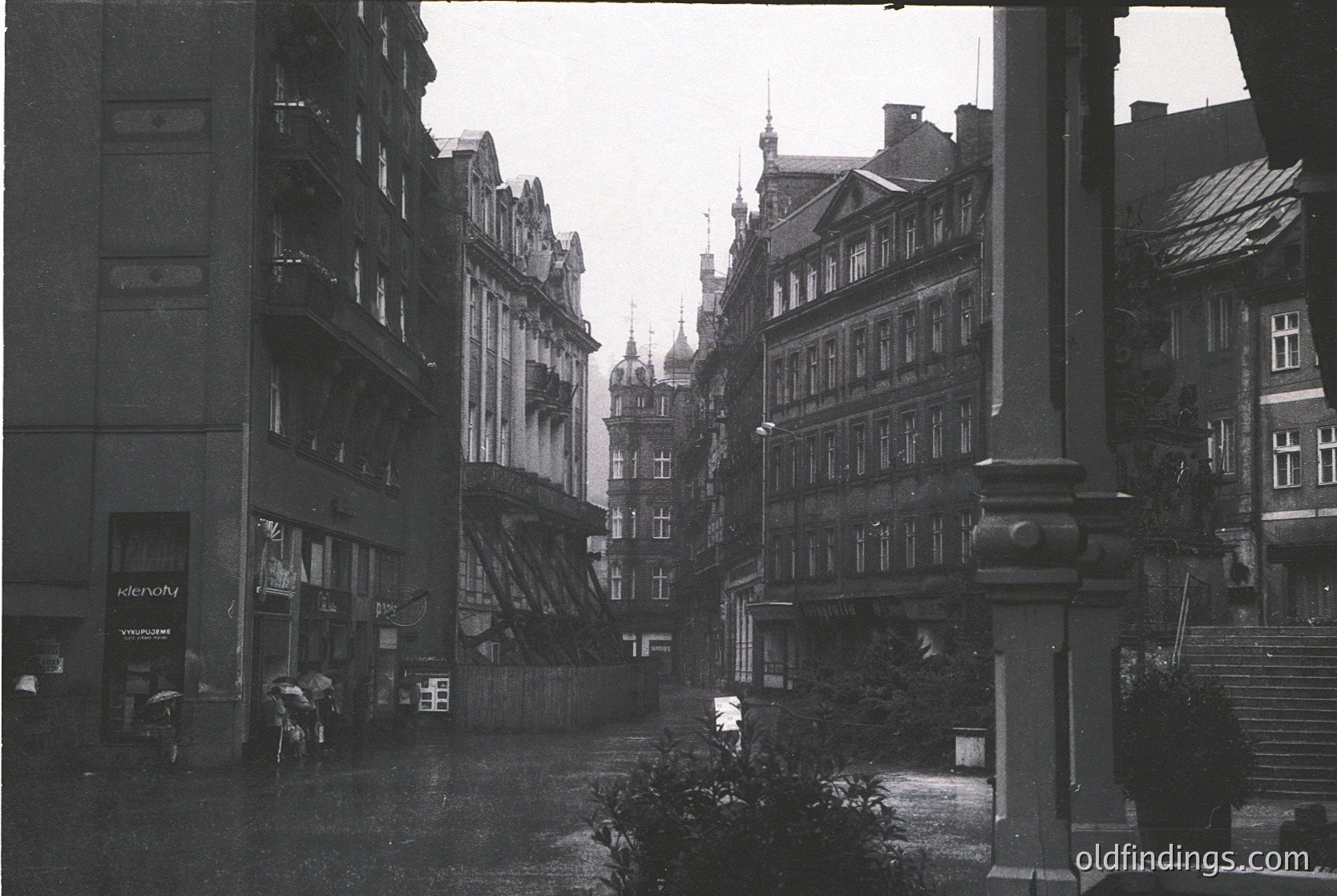 Mid-20th century European street scene featuring Gothic Revival architecture with steep gables, ornate facades, and wrought-iron balconies. Wet cobblestones reflect muted light; pedestrians carry umbrellas. Signage includes "Klenowy" and "Vinylux." Likely post-WWII reconstruction era.