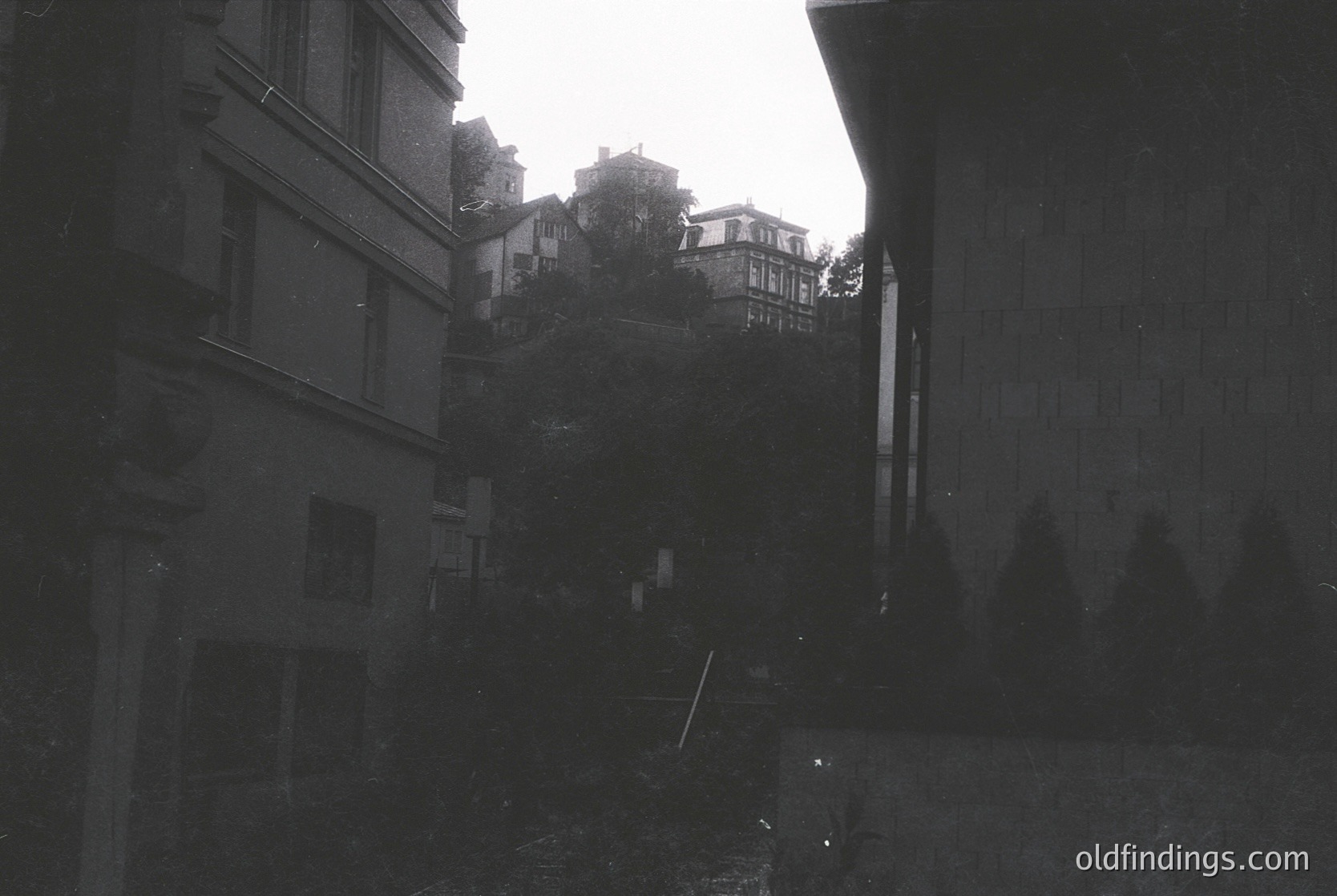 Black-and-white alleyway framed by tall, weathered buildings with exposed brick and concrete. Narrow pathway leads uphill to a hillside with scattered residential structures. Architectural style suggests mid-20th century European urban design. Rain-soaked pavement reflects muted light.
