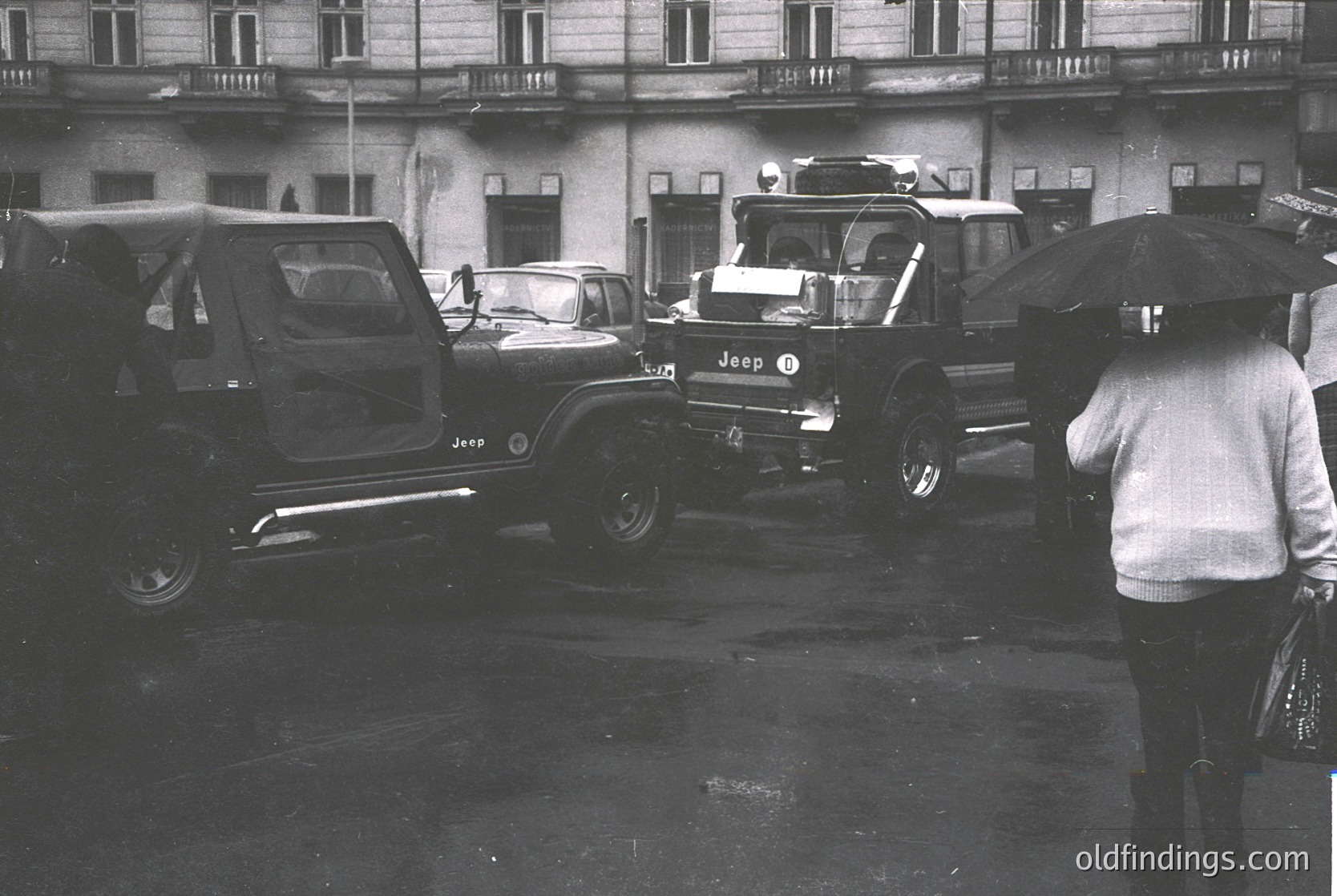 Vintage black-and-white street scene featuring a 1960s-era Jeep CJ-5 and other classic vehicles in a wet urban plaza. Multi-story building with balconies and large windows in background. Person holding umbrella, suggesting rain. Mid-century urban transport and architecture.