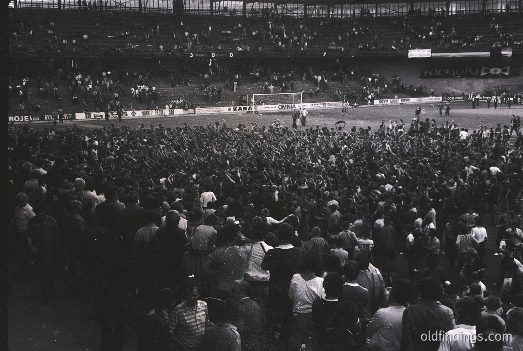 Black-and-white shot of a packed stadium during a football match, likely mid-20th century. Crowd fills stands, with visible scoreboard showing "3-0-0" and "SKRA" vs. "ROJEK." Advertisements for "KONSUM" and "SOKOL" on boards. Atmosphere captures mid-game intensity.