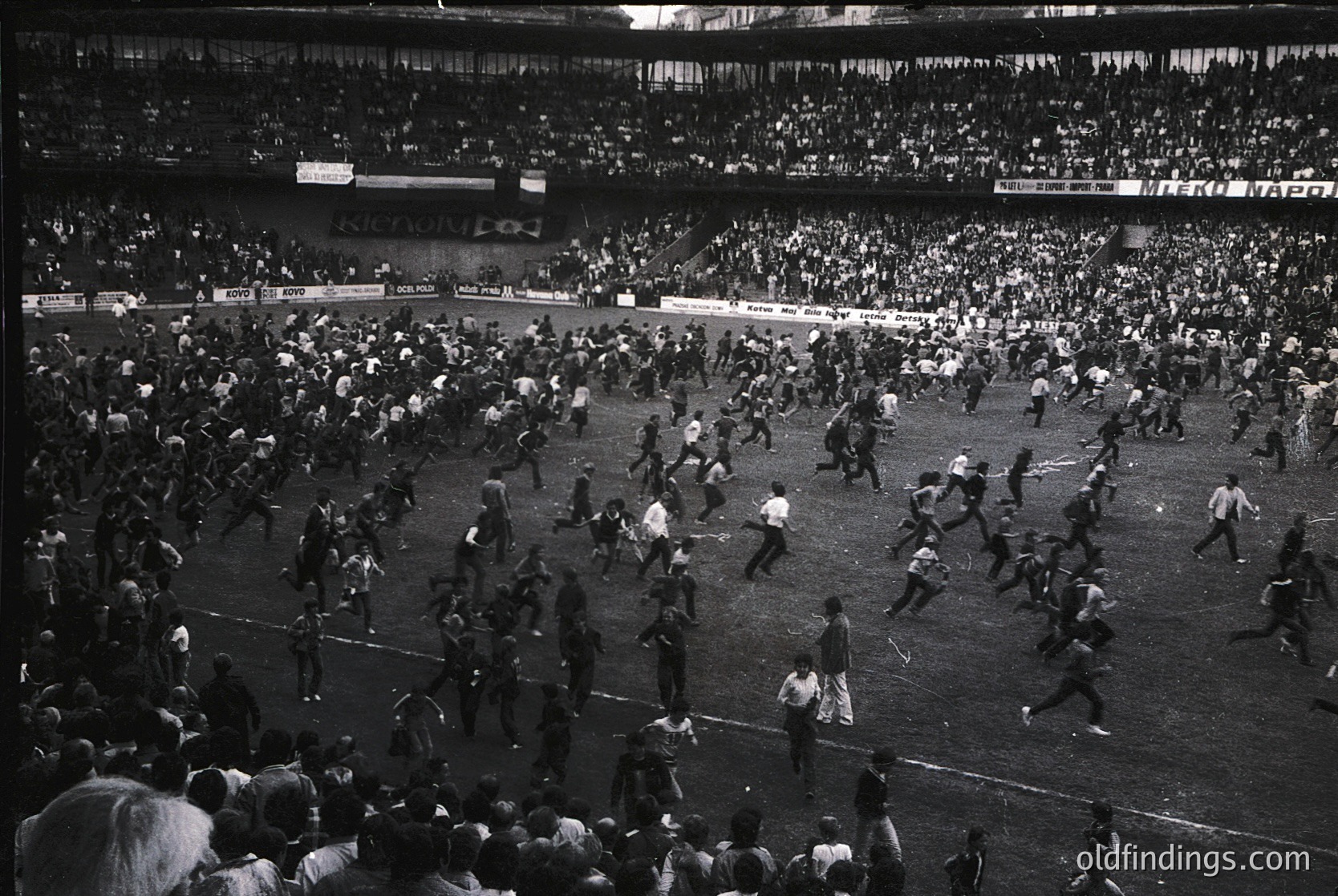 Chaotic stadium scene with players and fans flooding the field during a match. Mid-20th century stadium architecture with tiered seating and visible signage. Likely European football riot, 1960s–1980s era.