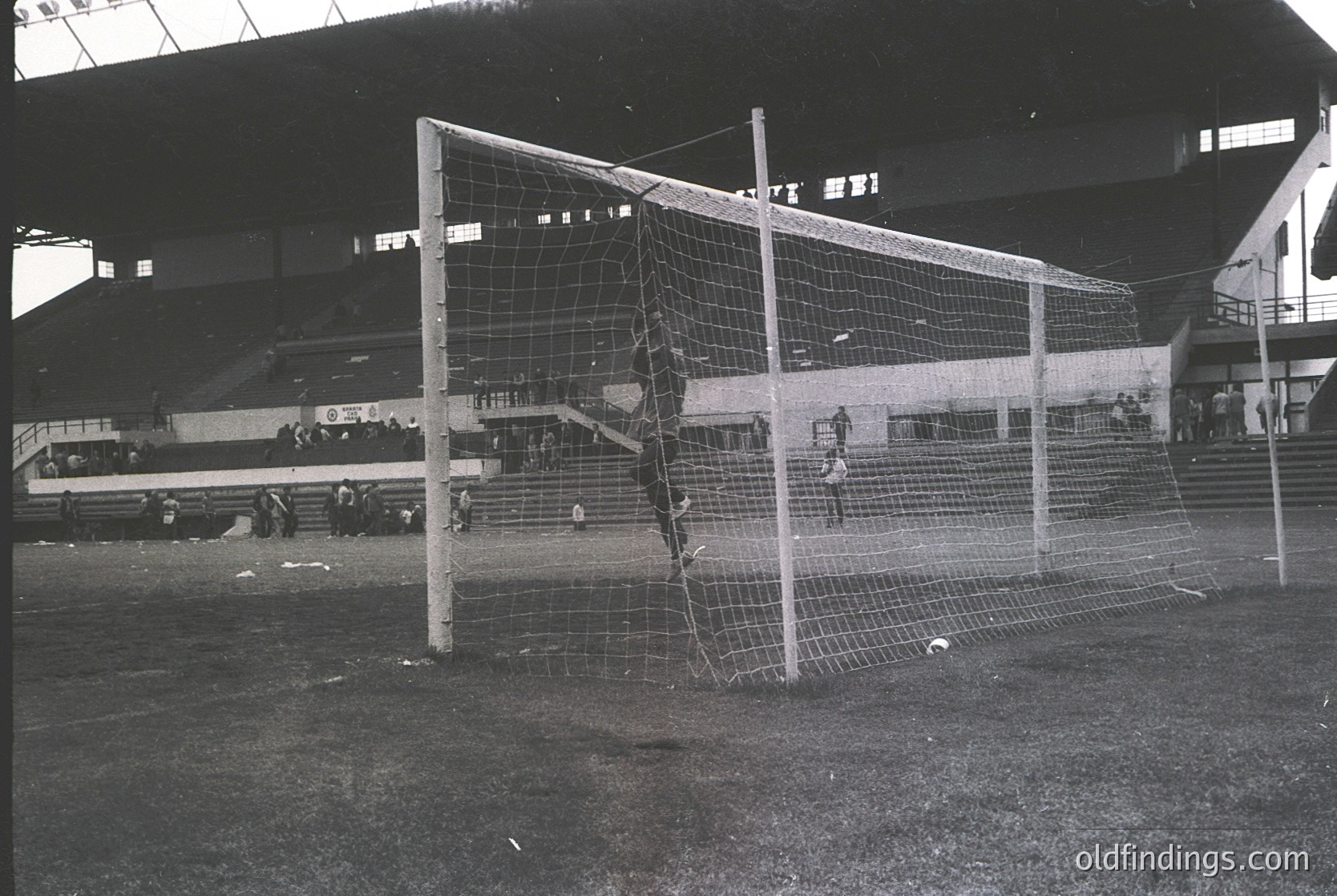 Empty mid-20th century stadium goalpost with netting damage, likely post-match. Concrete stands and sparse spectators in background suggest mid-century European design. Grass field and partial roof indicate an enclosed venue.