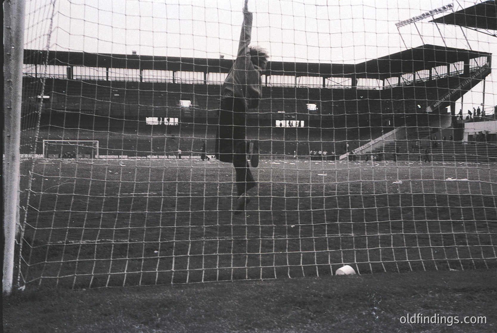 Black-and-white shot of a goalkeeper diving to block a soccer ball, mid-action. Empty stadium seats in background suggest a low-attendance or historical match. Classic net design and field markings indicate mid-20th century European football. Ideal for sports history or vintage sports photography.