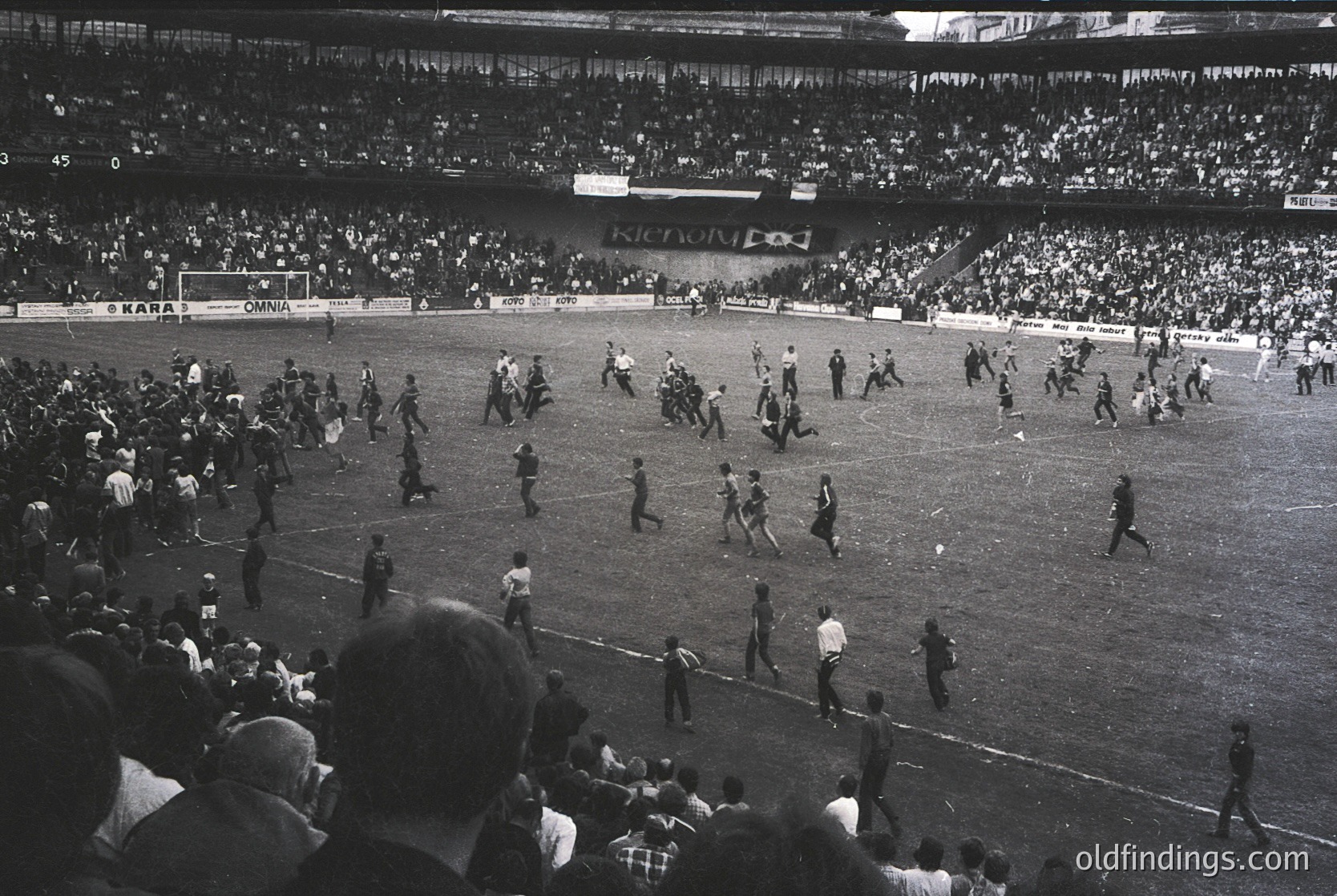 Black-and-white stadium shot showing chaotic post-match scene with players and officials scattered across the field. Crowd fills tiered stands, with visible advertisements like "KICOM" and "OKANA." Referees and players in uniform appear disoriented, likely during a 1960s–1970s European football match.