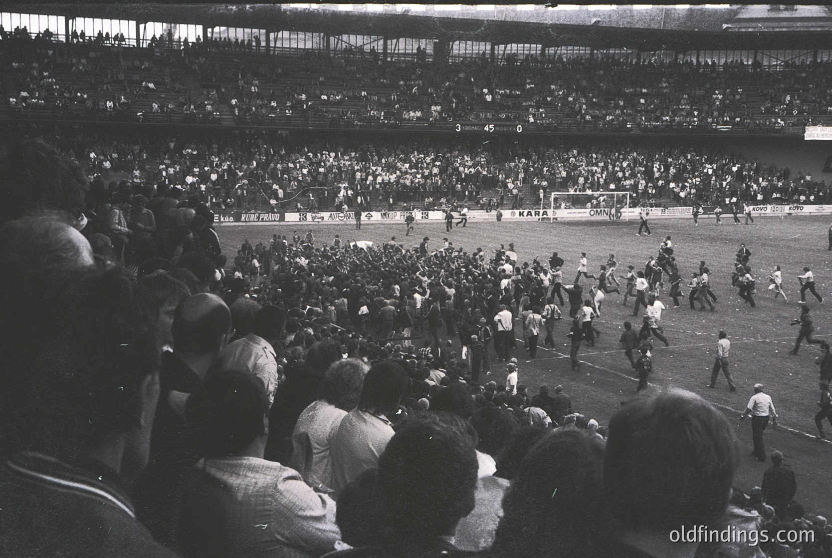 Vintage black-and-white stadium shot capturing a chaotic post-match scene, likely from the 1960s–70s. Crowd surges onto the field amid players and officials, with visible goalposts and a partially visible "UEFA" banner. High-energy atmosphere reflects mid-20th-century European football culture.