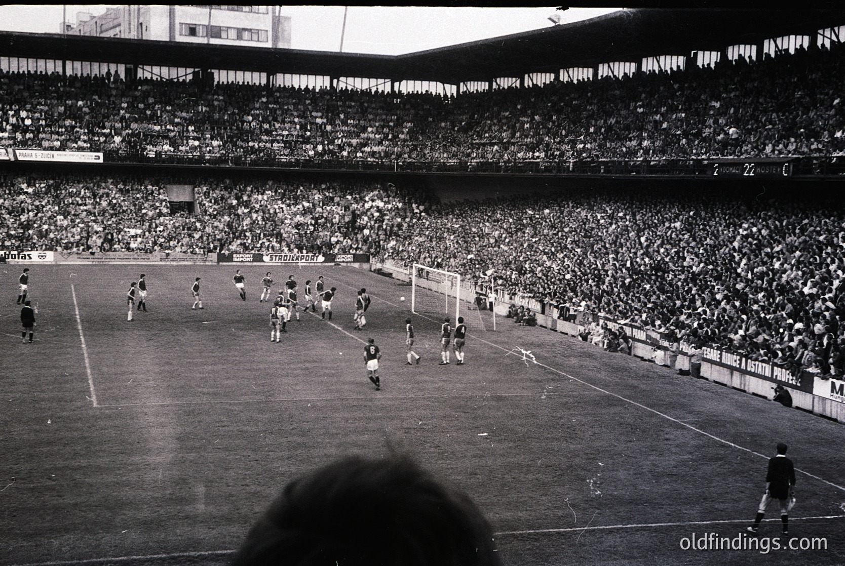 Vintage black-and-white shot of a packed stadium during a soccer match, likely 1960s–1970s. Crowd fills tiered stands, with players in dark uniforms near the goal. Architectural details include curved roofing and tiered seating. Iconic atmosphere of mid-20th-century European football.