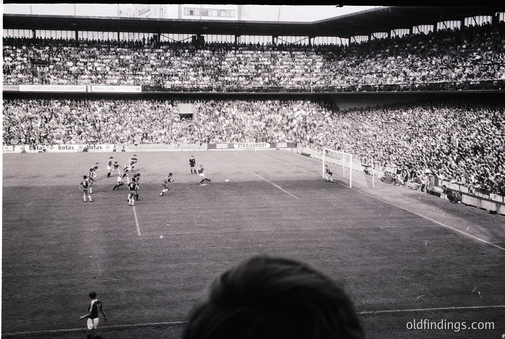 Black-and-white stadium shot captures a packed 1960s-era football match. Players in dark uniforms clash near the goal, while spectators fill tiered stands. Advertisements like "Botaş" and "Türk Hava Yolları" hint at Turkish setting. Crowd density and vintage grain evoke mid-century sports culture.