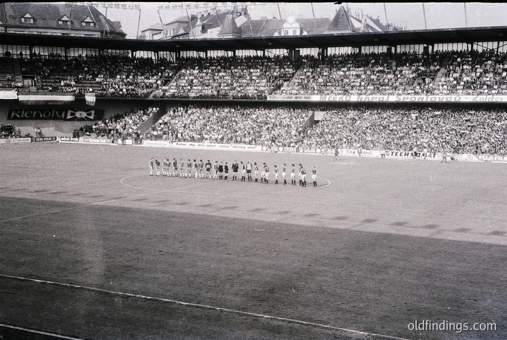 Black-and-white stadium photo showing a pre-game formation of a soccer team on a wet pitch, with a packed multi-tiered stand. Advertisement for "Klenol" visible on the upper tier. Crowd density suggests mid-20th century European match atmosphere.