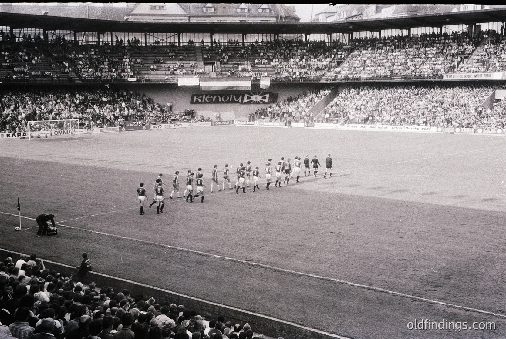 Black-and-white stadium photo showing pre-match team walkout on a grass pitch. Players in striped jerseys and shorts line up near the center circle, flanked by empty stands. Advertisements like "Kronospan" and "Kodak" visible on banners. Crowd fills lower tiers, indicating a mid-20th-century European football match. Crowded atmosphere suggests major league or cup event.