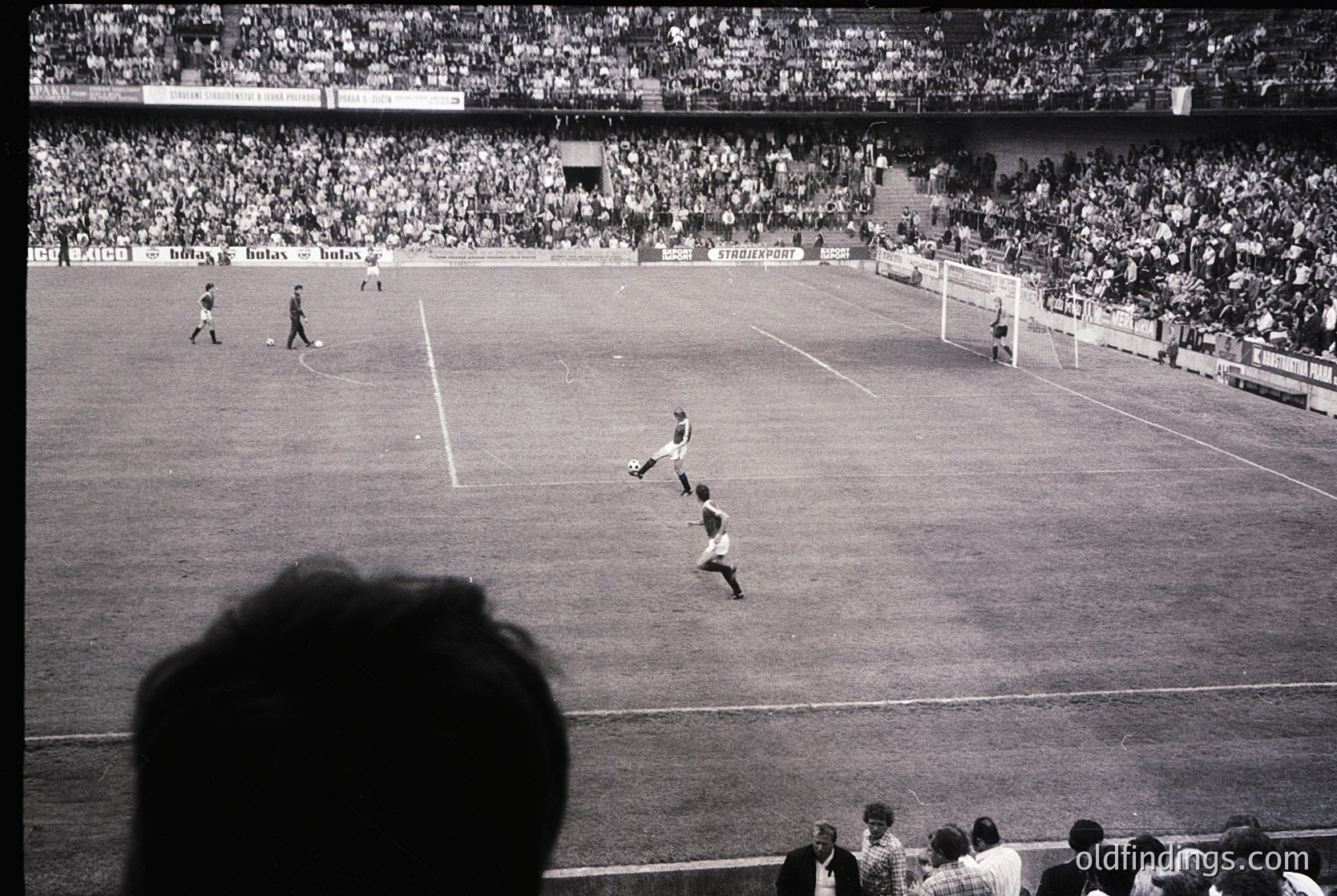 Black-and-white shot of a packed stadium during a mid-20th-century football match. A player in mid-air executing a header near the center of the pitch, with spectators densely filling stands. Advertising banners for brands like *Bulox* and *Stroopwafel* visible. Classic stadium architecture with tiered seating and open-air design.