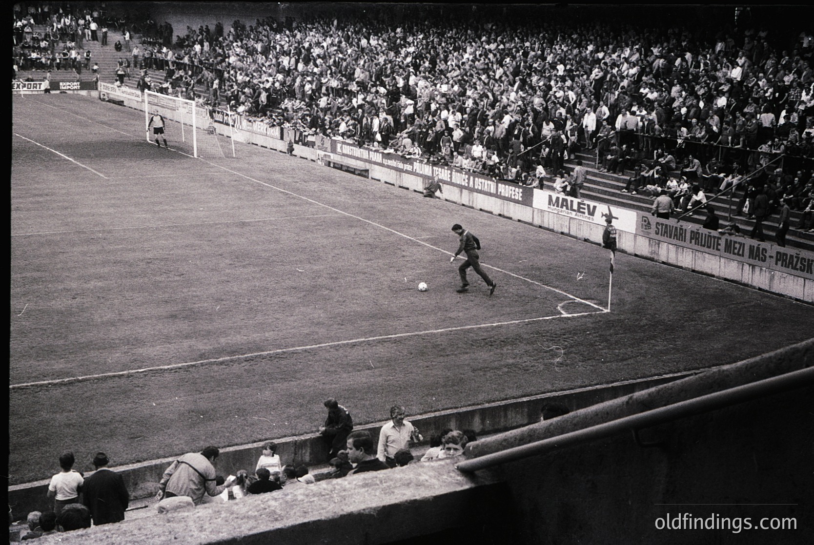 Black-and-white shot of a packed stadium during a soccer match, likely 1960s–1970s. Player in motion near the center circle, surrounded by spectators in tiered stands. Advertisements for "Malev" and "Stavari" visible on banners. Crowd density and stadium layout suggest European setting.