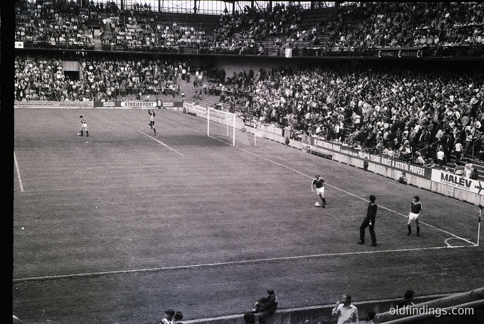 Black-and-white stadium shot capturing a 1960s–1970s football match. Crowded stands filled with spectators, visible branding like "Marlboro" and "Tiger" ads. Players in vintage striped jerseys and shorts, referee in dark uniform. Grass field with goalposts and corner flags. Iconic mid-match energy.