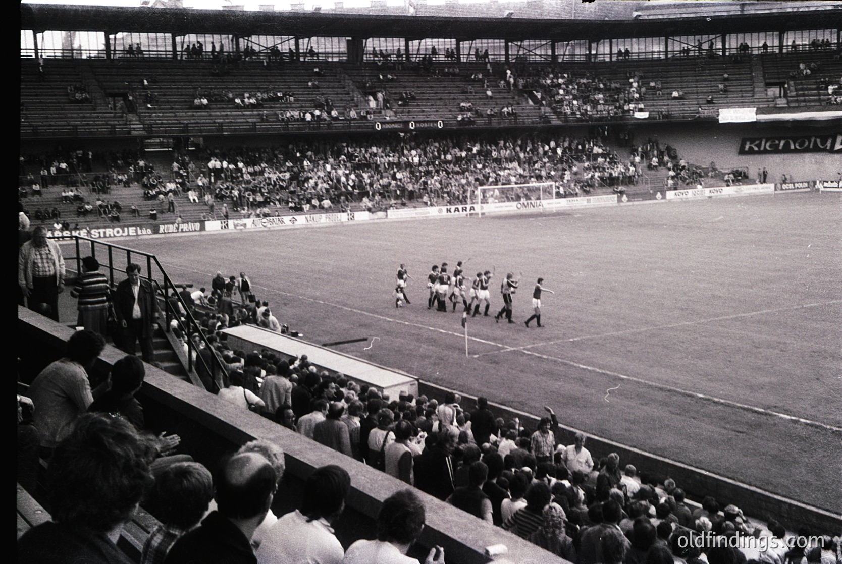 Black-and-white stadium shot showing a mid-20th-century football match. Players in vintage striped jerseys line up for kickoff on a grass pitch, with spectators densely packed in stands. Advertisements for brands like Ricoh and Karpi are visible. Architectural details include tiered seating and a covered press box.