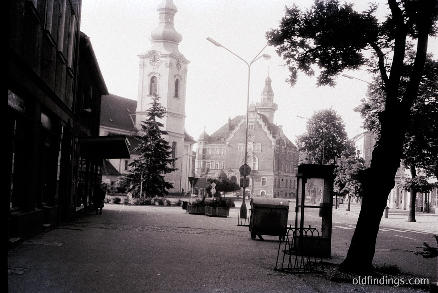 Black-and-white street scene featuring **1960s-70s European urban architecture**. Prominent **baroque-style church tower** with clock and ornate facade dominates center. Adjacent **historic building** with mansard roof and decorative gables. Empty **street lamp**, **street sign**, and **public phone booth** suggest quiet era. Lush **street trees** and **planters** add greenery. Likely **Central/Eastern Europe** due to architectural style.