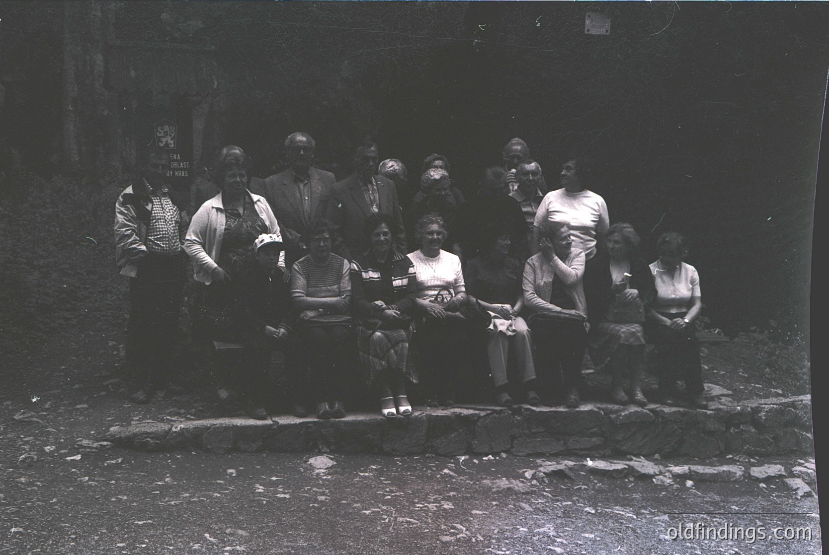 Black-and-white group portrait of 12 individuals seated on a low stone wall, likely mid-20th century. Formal attire includes suits, dresses, and headscarves; one child in a striped shirt. Dark, textured background suggests an urban or industrial setting.