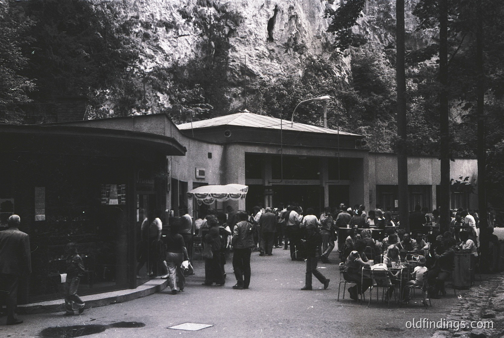 Mid-century urban courtyard with mid-rise building featuring flat roof and minimalist architecture. Crowd of casually dressed people—some seated at tables, others walking—suggests a public gathering or event. Decorative garlands indicate a festive occasion. Lush trees frame the scene, hinting at a European cityscape. Likely 1950s–1960s.