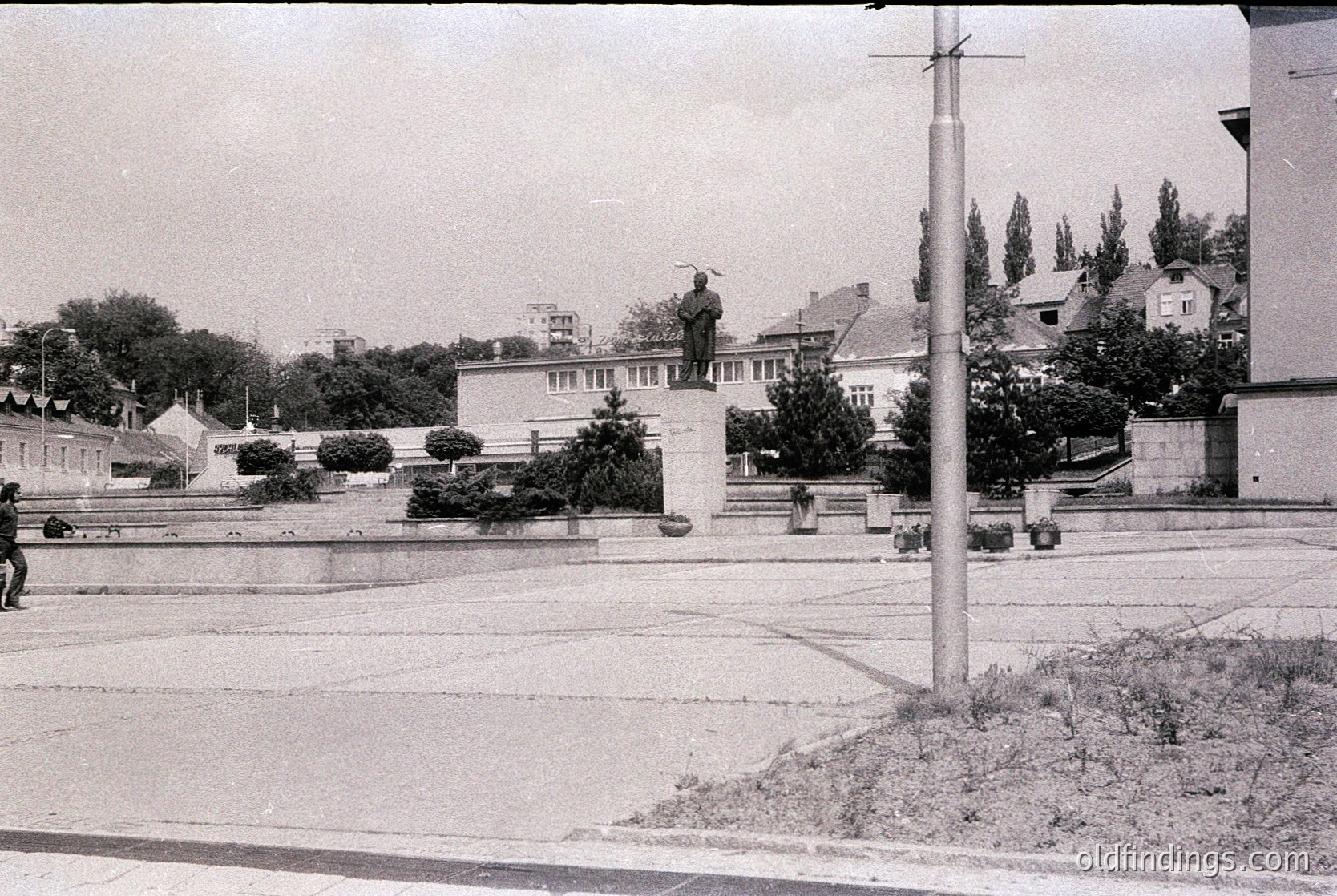 Monumental statue atop pedestal in urban plaza, flanked by low-lying Soviet-style architecture. Prominent tree-lined street with utility poles and sparse pedestrians. Likely Eastern Bloc urban design, mid-20th century.