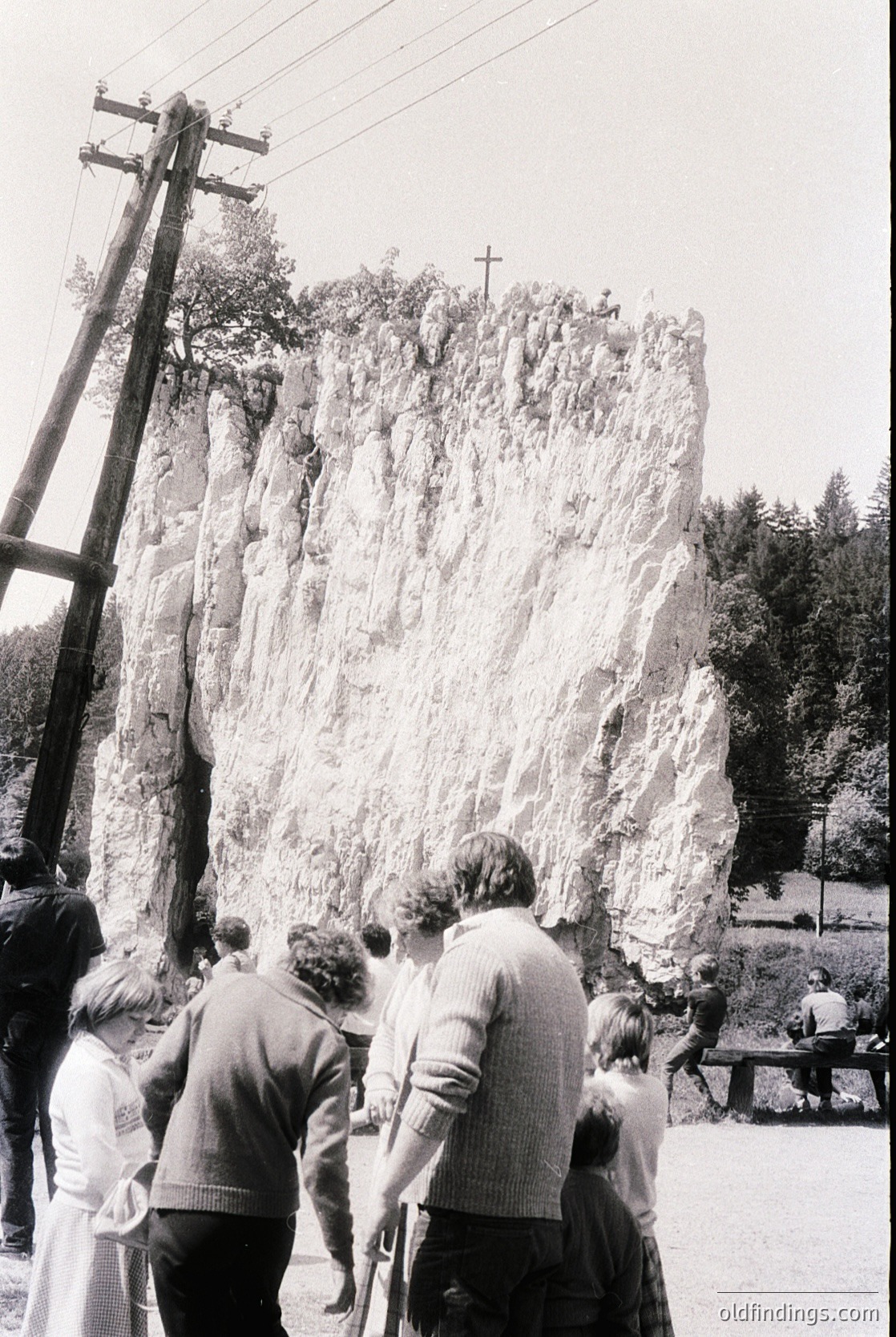 Mid-20th century group of people observing a large, weathered rock formation with a cross carved at its summit. Clothing suggests or Eastern European setting. Urban park or public space with utility poles and paved area. Potential religious or cultural pilgrimage site.