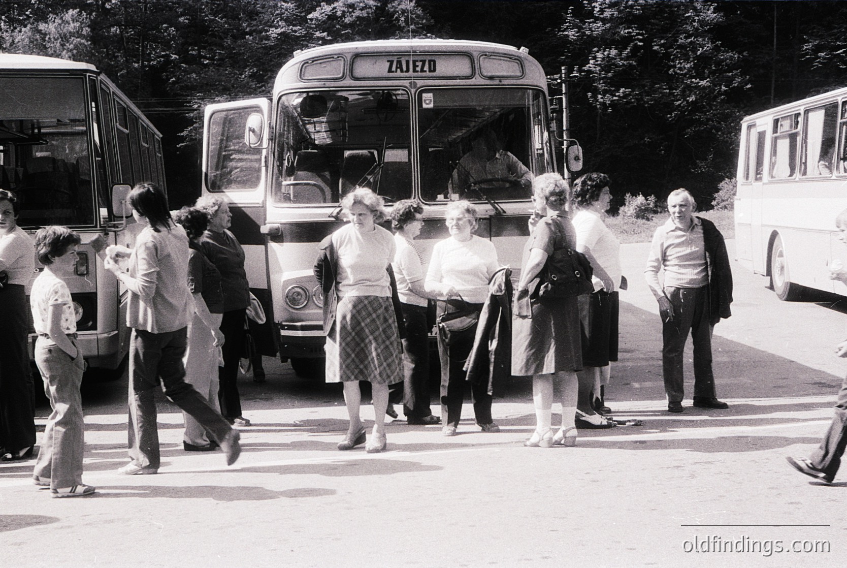 A group of people boarding a vintage **Zajezd** bus (likely Eastern European, mid-20th century) in an outdoor setting. Casual attire suggests a 1960s–1970s timeframe. Trees and greenery frame the scene, indicating a suburban or park area.