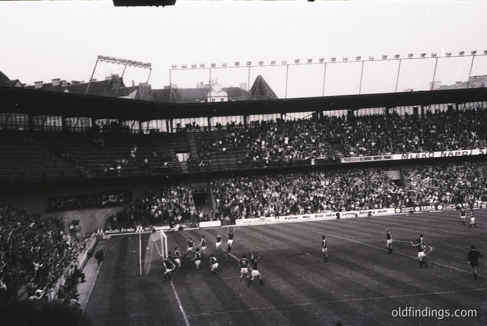 Black-and-white shot of a packed stadium during a mid-20th-century football match, likely 1950s–1960s. Crowds fill tiered stands, with players in white and dark uniforms near the goal. Floodlights and advertising banners (e.g., "NAVITAR") indicate a professional venue. Architectural details suggest European design.