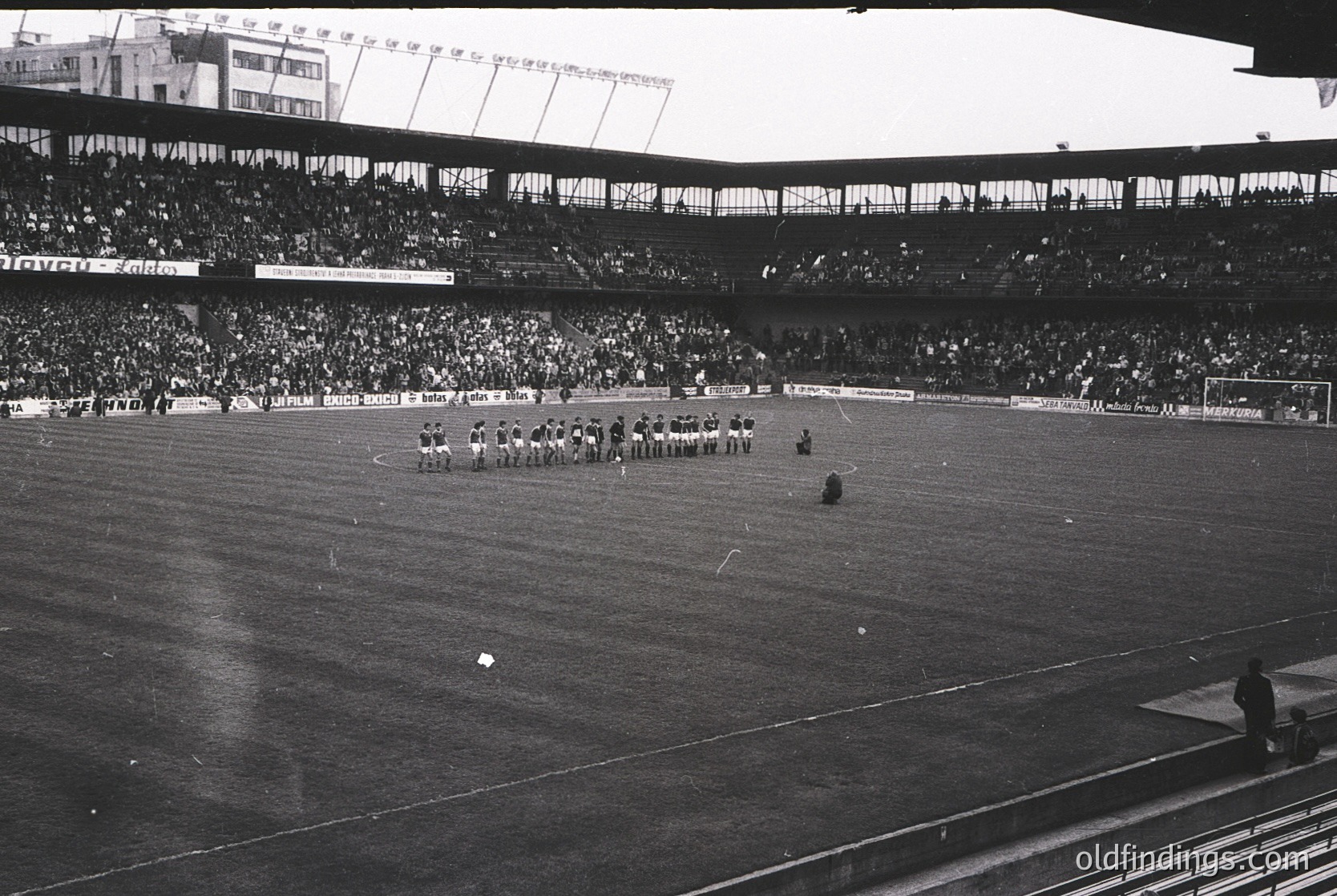 Vintage black-and-white stadium shot showing a midfield kickoff with players in formation. Crowded stands with tiered seating and visible advertisements (, ). Classic American stadium architecture with floodlights and open-air design.