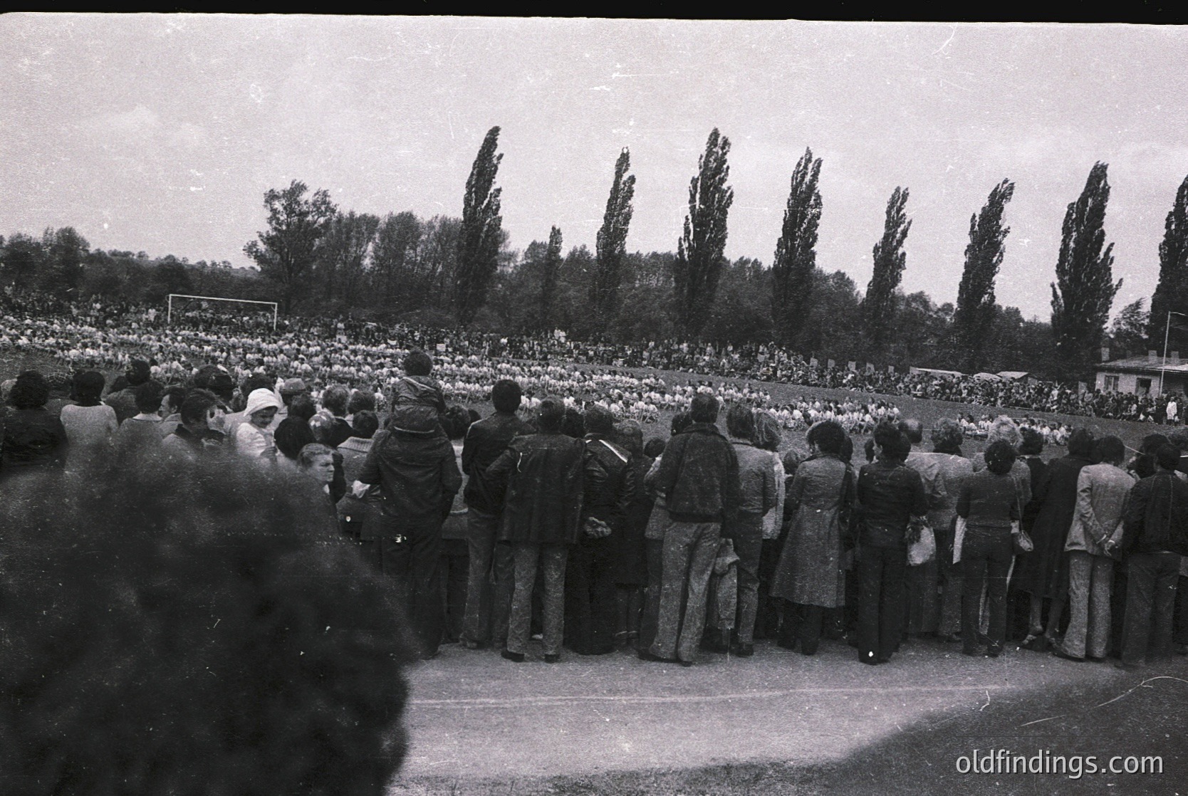 Black-and-white stadium shot of dense crowd facing a soccer field, likely mid-20th century. Uniform attire suggests organized event—potential sports match or public gathering. Trees frame the background, indicating outdoor venue.