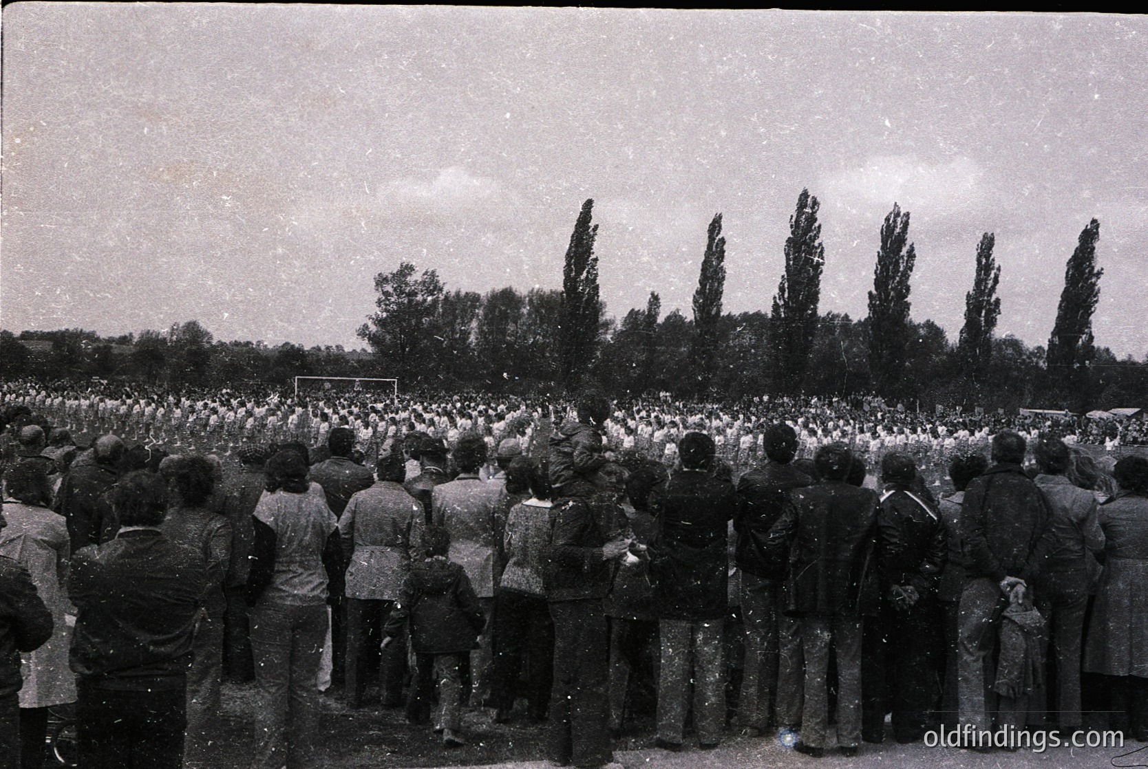 Crowd of people gathered outdoors in a formal, organized procession, likely a funeral or memorial. Uniform attire suggests a collective mourning event. Rows of trees and open field in background. Black-and-white, high-contrast photography suggests mid-20th century (1940s–1960s).