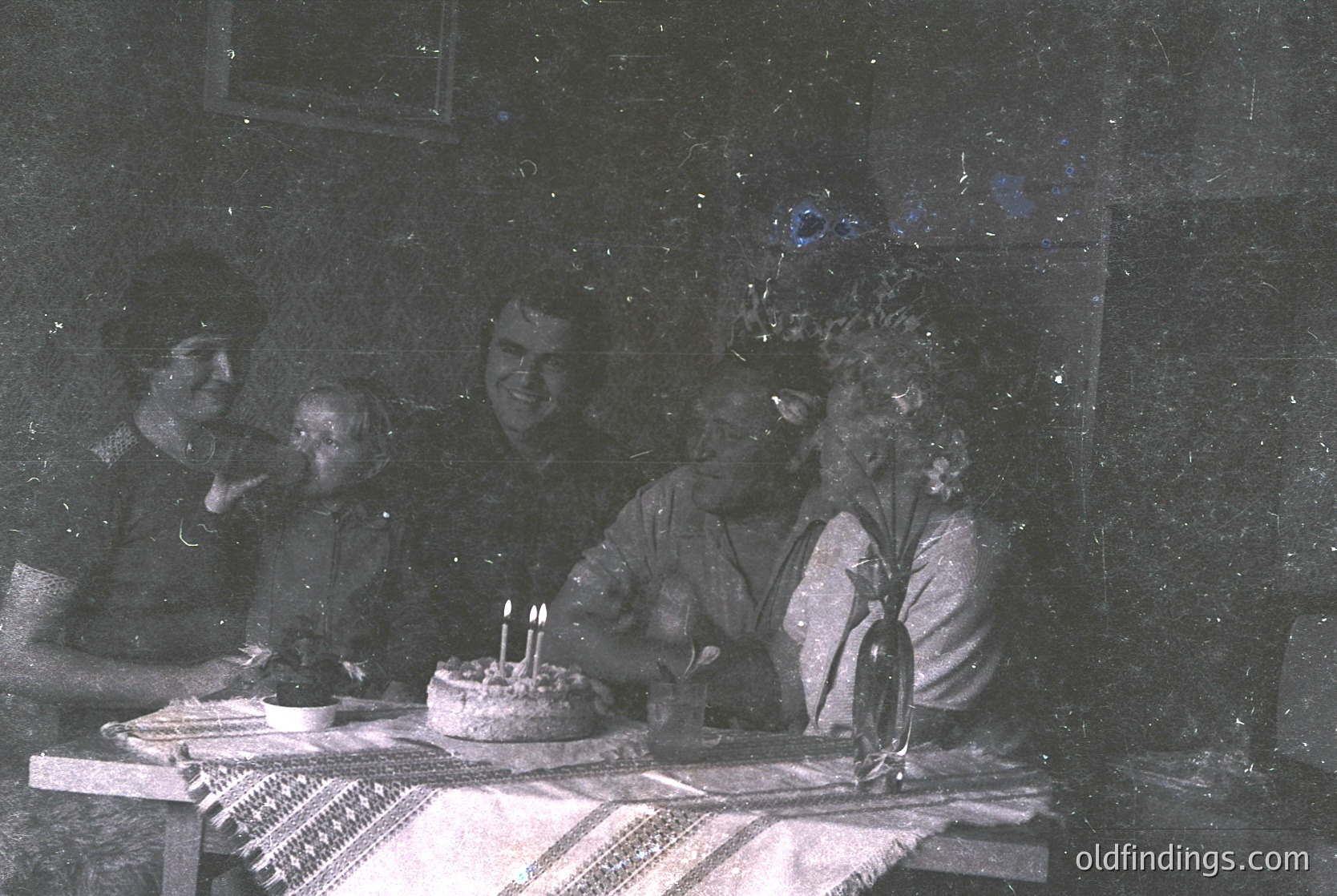 Vintage sepia-toned group portrait featuring five individuals seated around a table with a small cake and lit candles. Indoor setting with blurred background suggesting a dimly lit room. Traditional patterned tablecloth and simple tableware indicate mid-20th century domestic celebration.