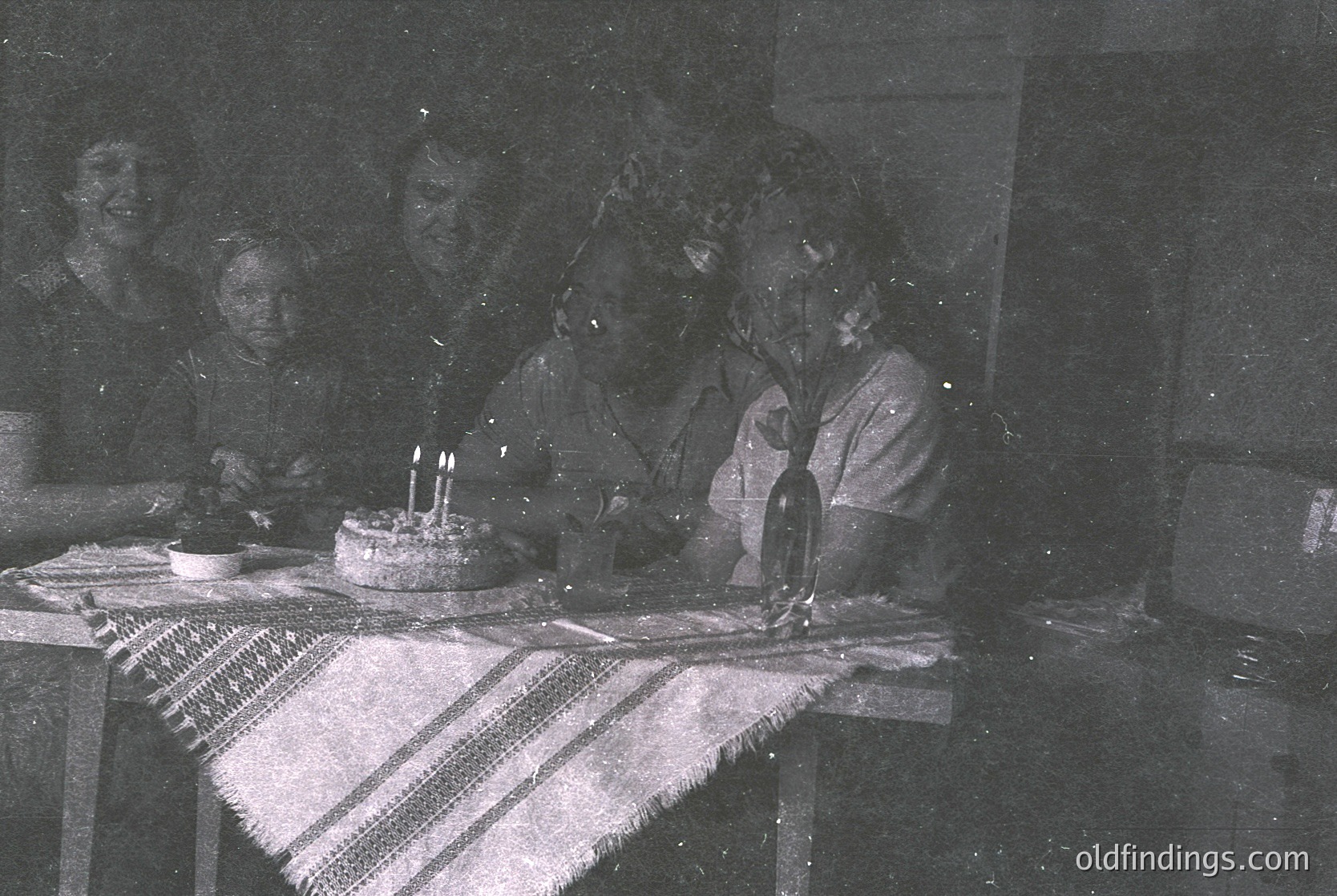 Vintage black-and-white indoor celebration featuring a woman blowing out candles on a simple round cake. Surrounding her are blurred figures seated at a table draped with a striped cloth. Bottle and plates suggest a modest gathering, likely mid-20th century.