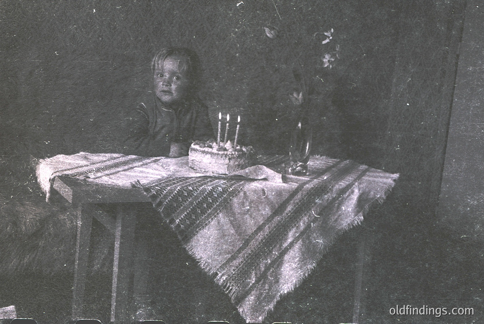 Vintage sepia-toned photo of a young child seated at a small wooden table covered with a striped cloth, blowing out birthday candles on a cake. Indoor setting with minimal lighting, likely mid-20th century.
