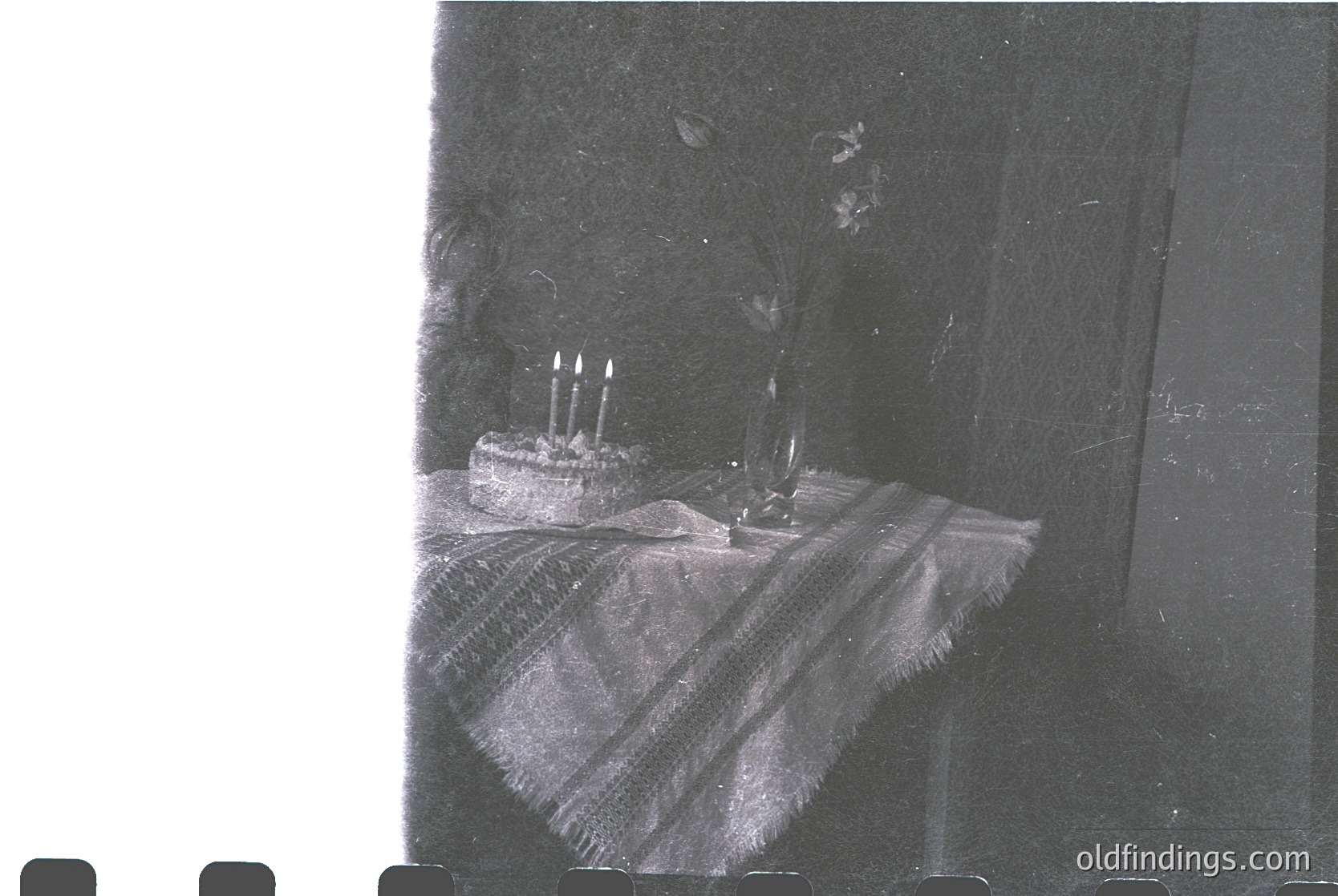 Vintage black-and-white photo of a simple birthday cake with three lit candles on a striped tablecloth, likely 1950s-1970s. Indoor setting with minimalist decor, suggesting a modest celebration.