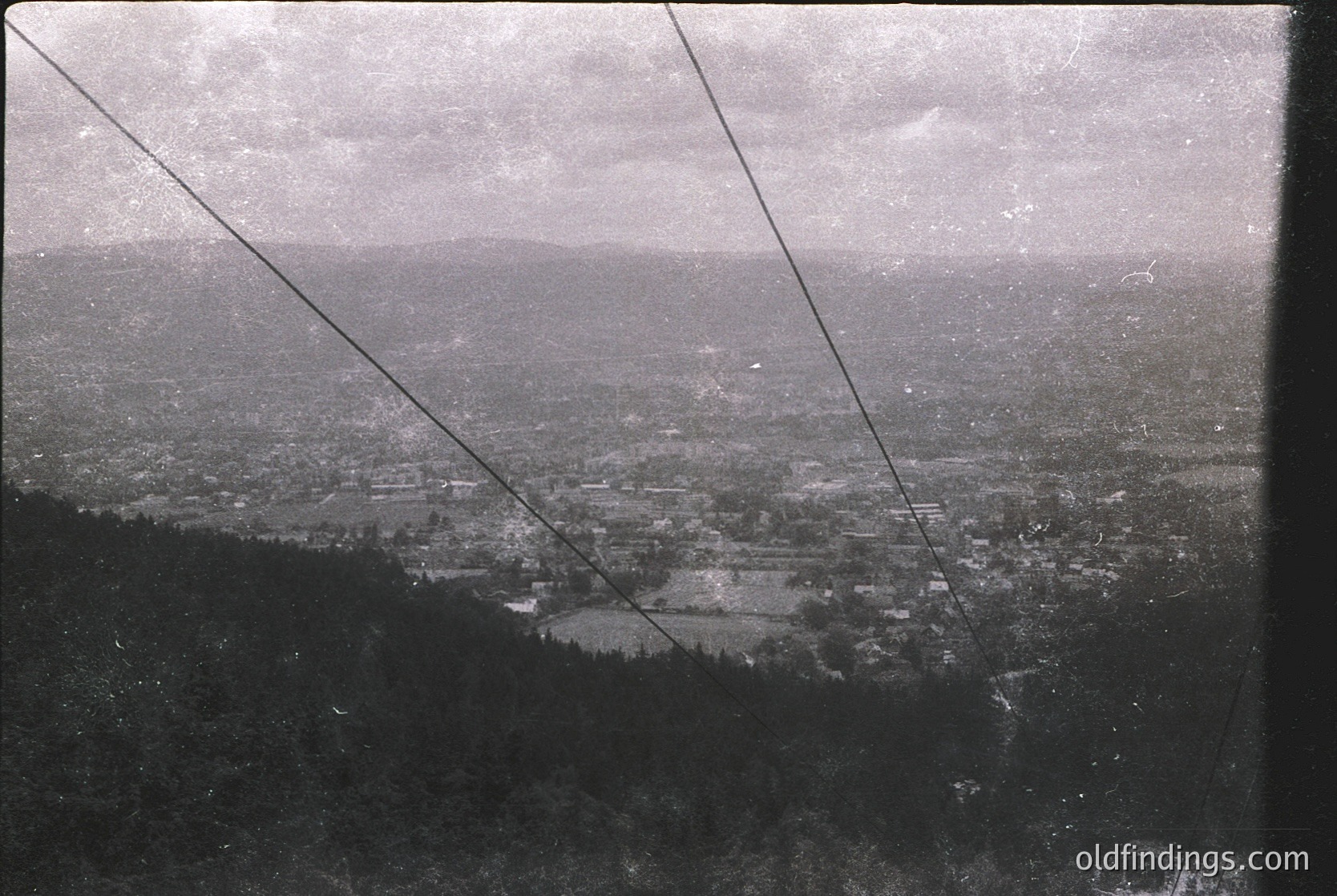 Vintage aerial view of a sprawling urban landscape, likely captured via early 20th-century photography. Dense grid of buildings, roads, and greenery stretches toward distant hills under overcast skies. Cable lines frame the composition, suggesting a high vantage point. Urban planning and early industrial architecture visible.