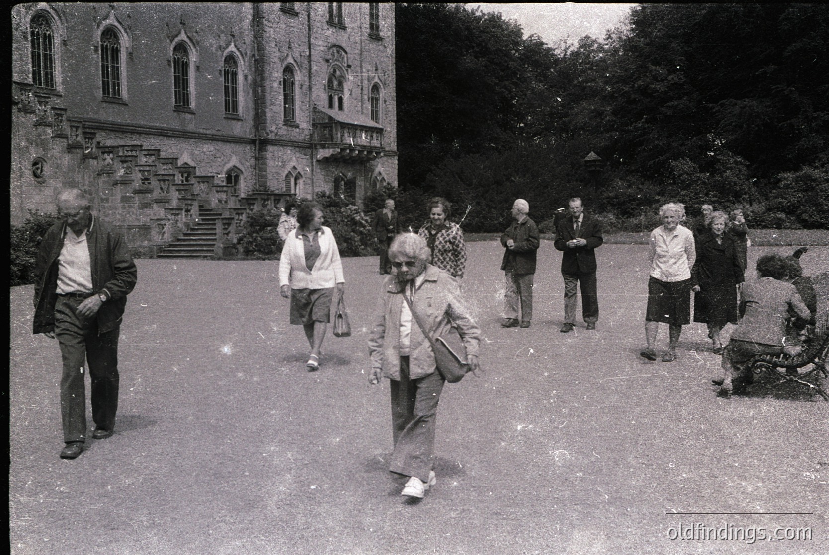 Gothic-style building with pointed arches and stone detailing, mid-20th century. Group of elderly individuals in casual 1960s-70s attire—men in suits, women in dresses/blazers—walking and standing in a landscaped courtyard. Lush greenery and mature trees frame the scene.