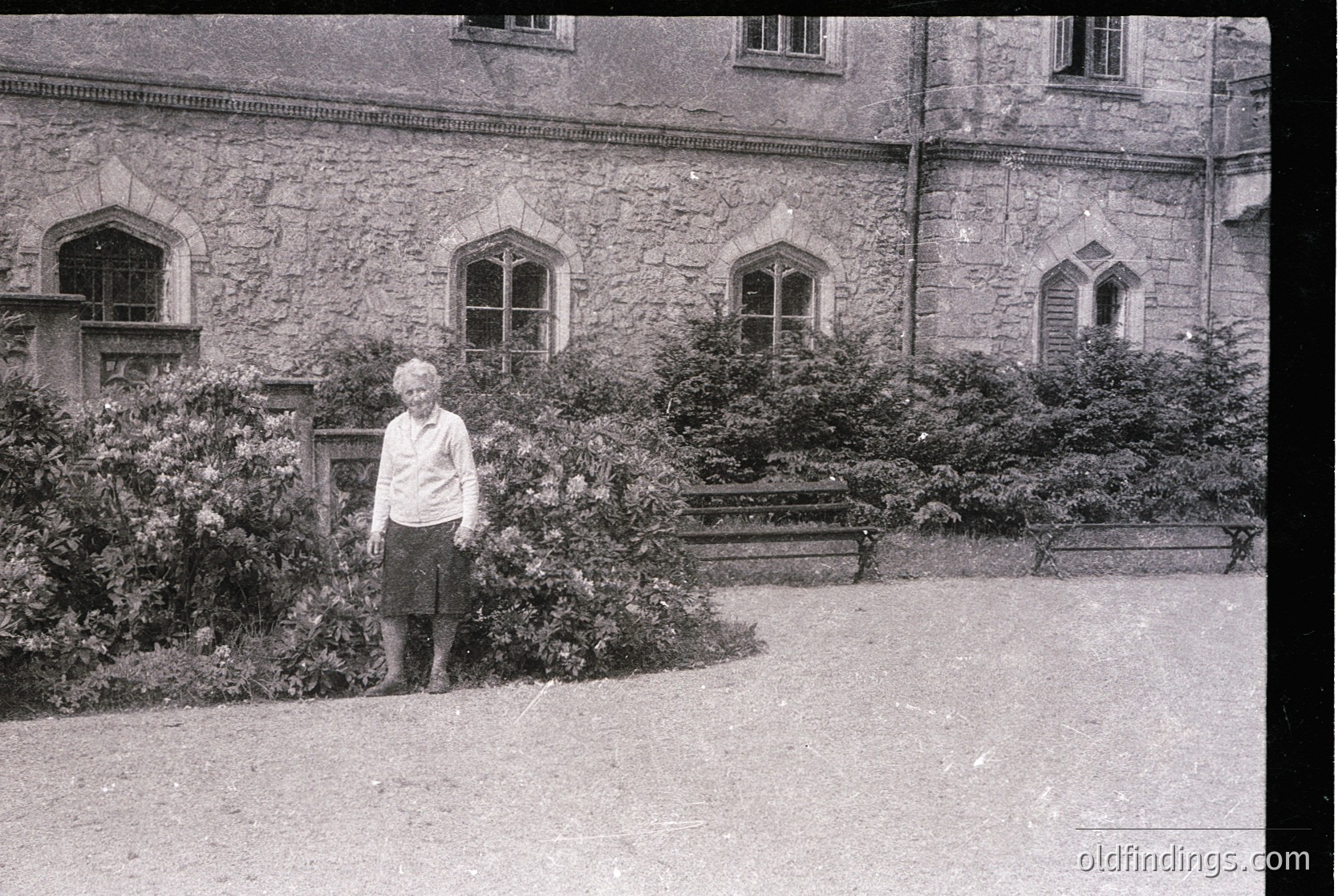 Vintage black-and-white portrait of an elderly woman standing beside a stone building with arched windows, surrounded by dense shrubbery. Mid-20th century attire suggests fashion. Courtyard setting evokes historical European architecture.