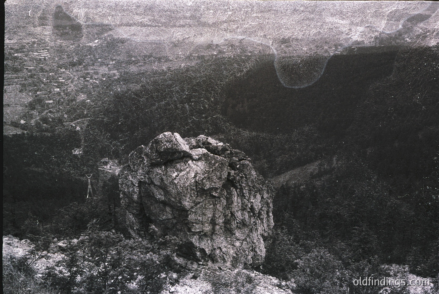 Aerial black-and-white photo of a coastal rock formation jutting into the sea, likely taken during mid-20th century. The jagged rock contrasts with the smooth, dark water below, while faint urban structures and roads are visible in the background.