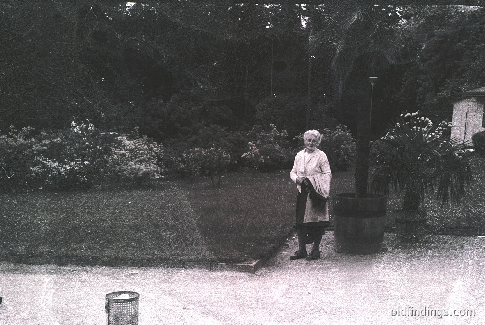 A mid-20th century woman in a light coat and dark skirt stands in a landscaped garden, holding a handbag. Lush greenery, including palm trees and shrubs, surrounds a paved pathway. The sepia tone suggests vintage photography, likely or .
