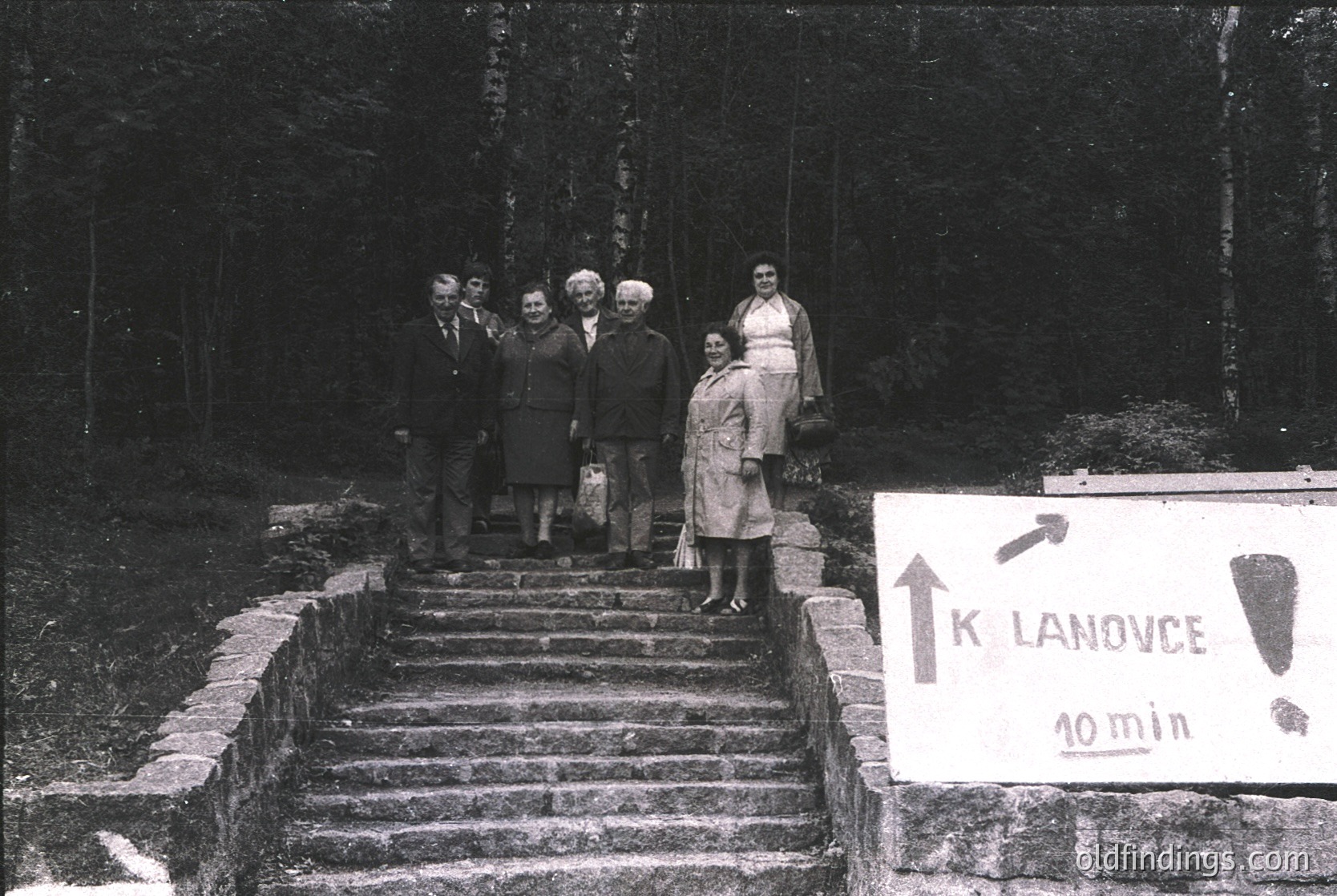 Black-and-white group photo on stone steps leading to a hand-painted wooden sign reading *"K LANOVCE 10 min"* in Czech, indicating a ski lift destination. Seven adults pose formally, dressed in 1960s-70s mountainwear—coats, cardigans, and hats. Forest backdrop suggests alpine terrain.