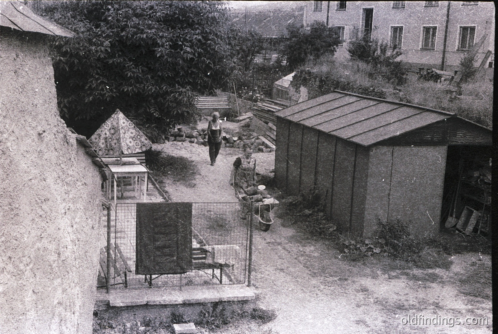 Aerial view of a courtyard with mid-20th-century urban housing. Concrete walls, metal-fenced utility area with a small cage-like structure (likely a pigeon loft or ventilation shaft) and a metal shed. Two individuals seated on concrete steps, one near a small table with a covered object. Lush greenery contrasts with the utilitarian architecture. Likely Eastern Bloc-era residential block.
