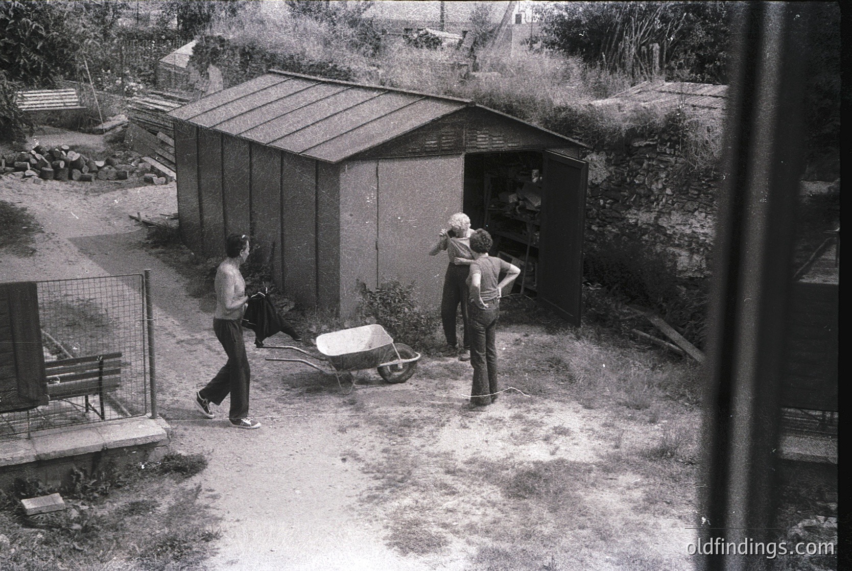 Mid-20th-century backyard scene with two men in casual workwear (bell-bottoms, button-ups) near a corrugated metal shed. One man pushes a wheelbarrow; the other stands near a fence. Concrete slab and gravel ground suggest construction or gardening activity.