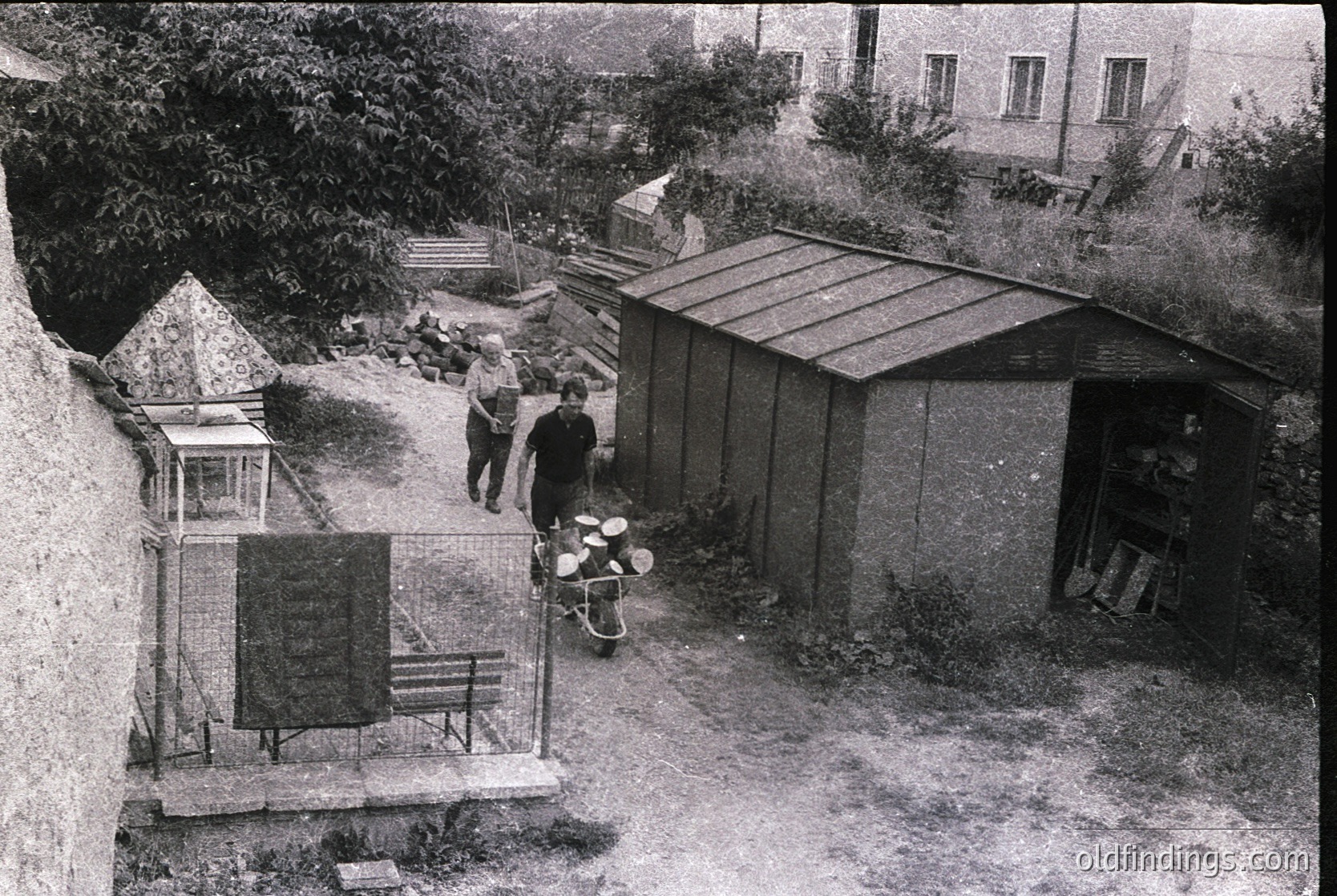 Aerial view of a mid-20th-century courtyard with Soviet-era architecture. Three individuals stand near a metal bench and a small wooden shed, while a bicycle leans against a fence. A Soviet flag and a decorative pyramid structure with Cyrillic lettering (likely Bulgarian) are visible. Overgrown greenery and a residential building frame the scene.