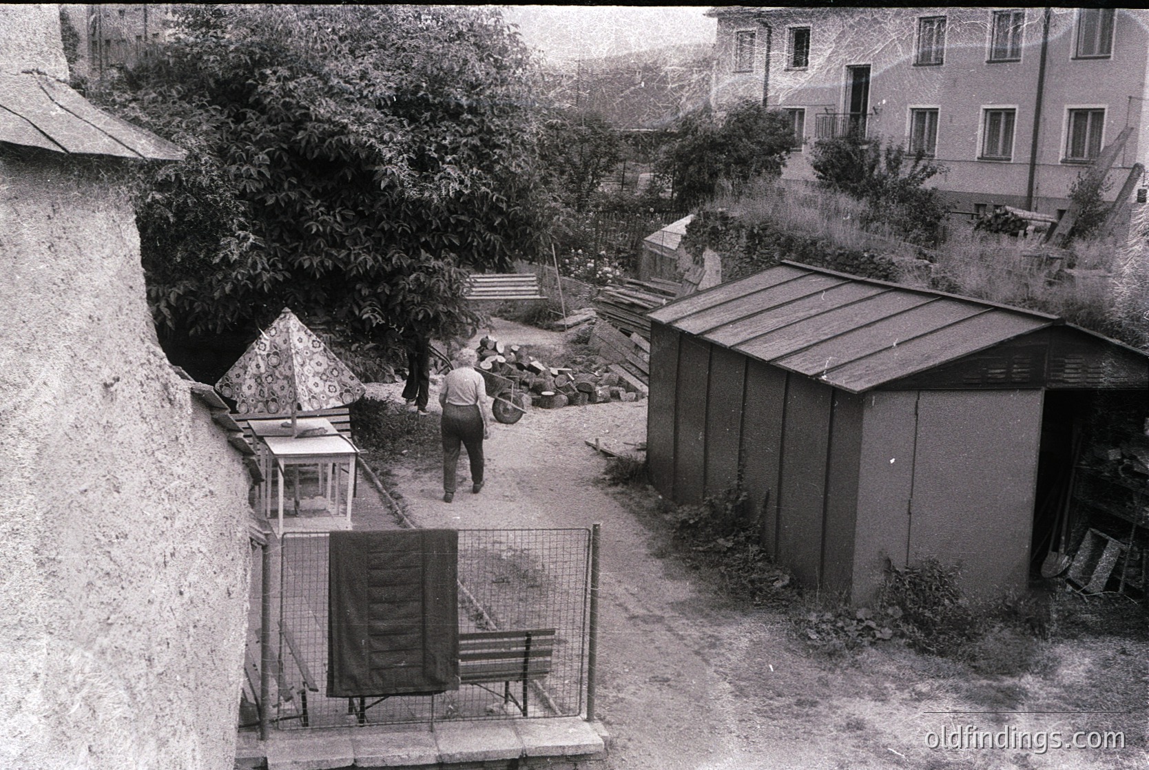 Vintage courtyard scene with concrete walls, metal fencing, and utilitarian structures. A man in mid-step walks toward a small shed, while a birdcage and wooden bench sit near a stone wall. Mid-century residential architecture in background. Likely Eastern Europe, 1960s-70s.