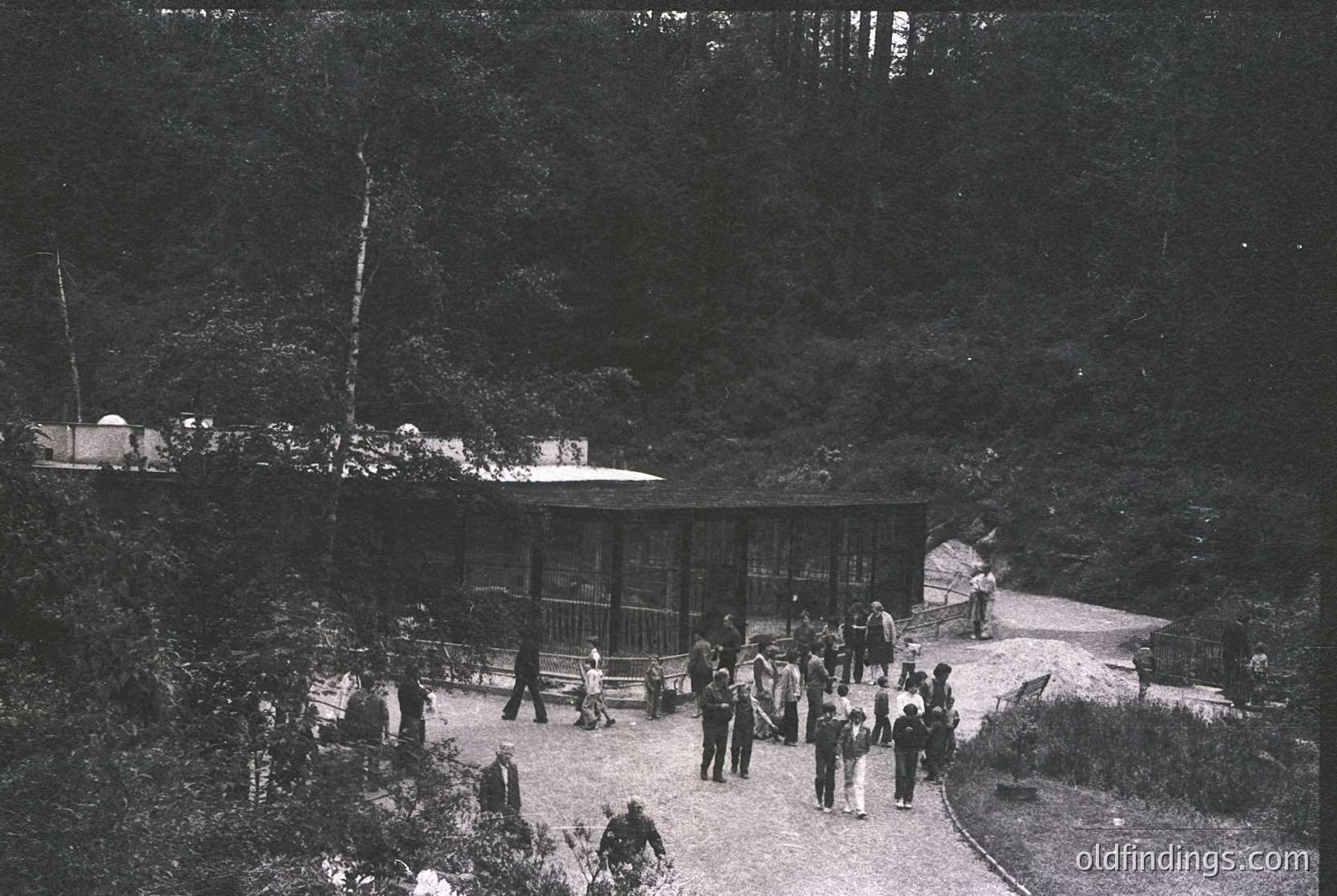 A vintage black-and-white photo of a forested hillside resort with a wooden pavilion and open-air seating. Mid-20th century attire suggests 1940s–1950s era. Crowd gathers near a curved pathway, hinting at social or recreational gathering.