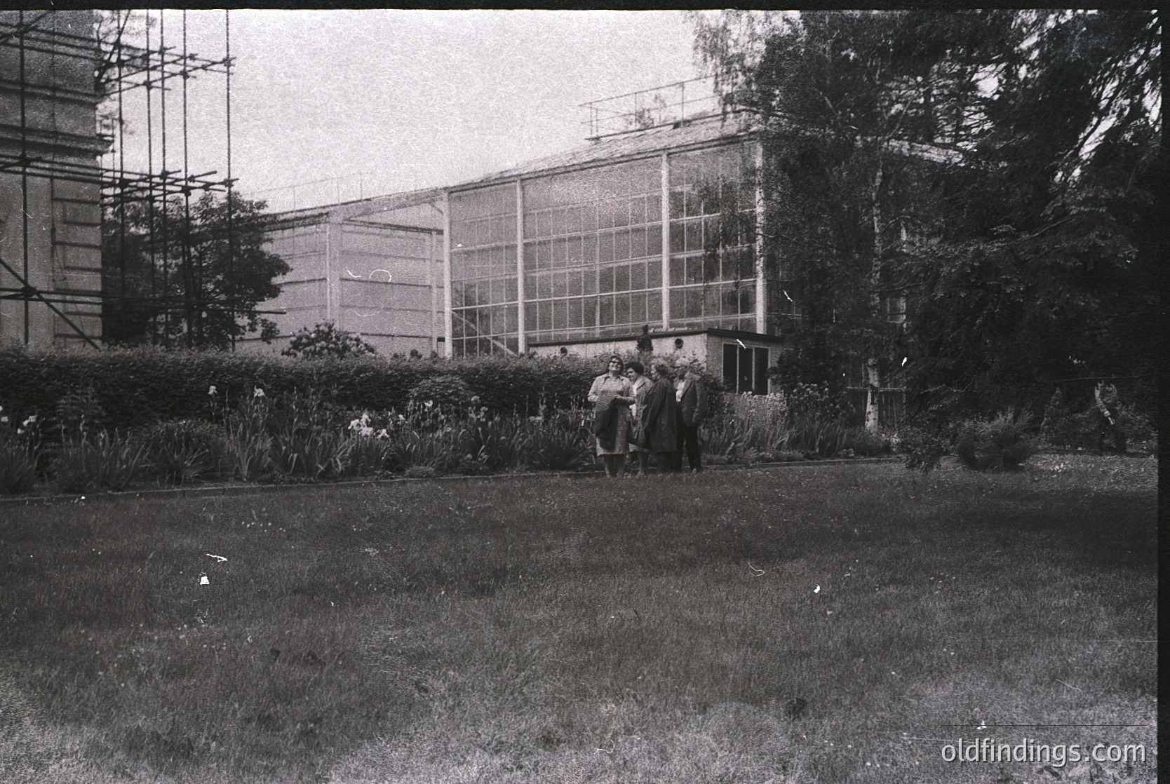 Mid-20th century botanical conservatory with large glass panels and scaffolding, likely 1950s-60s. Group of people in formal attire poses near manicured garden beds. Architectural detail reflects post-war institutional design.