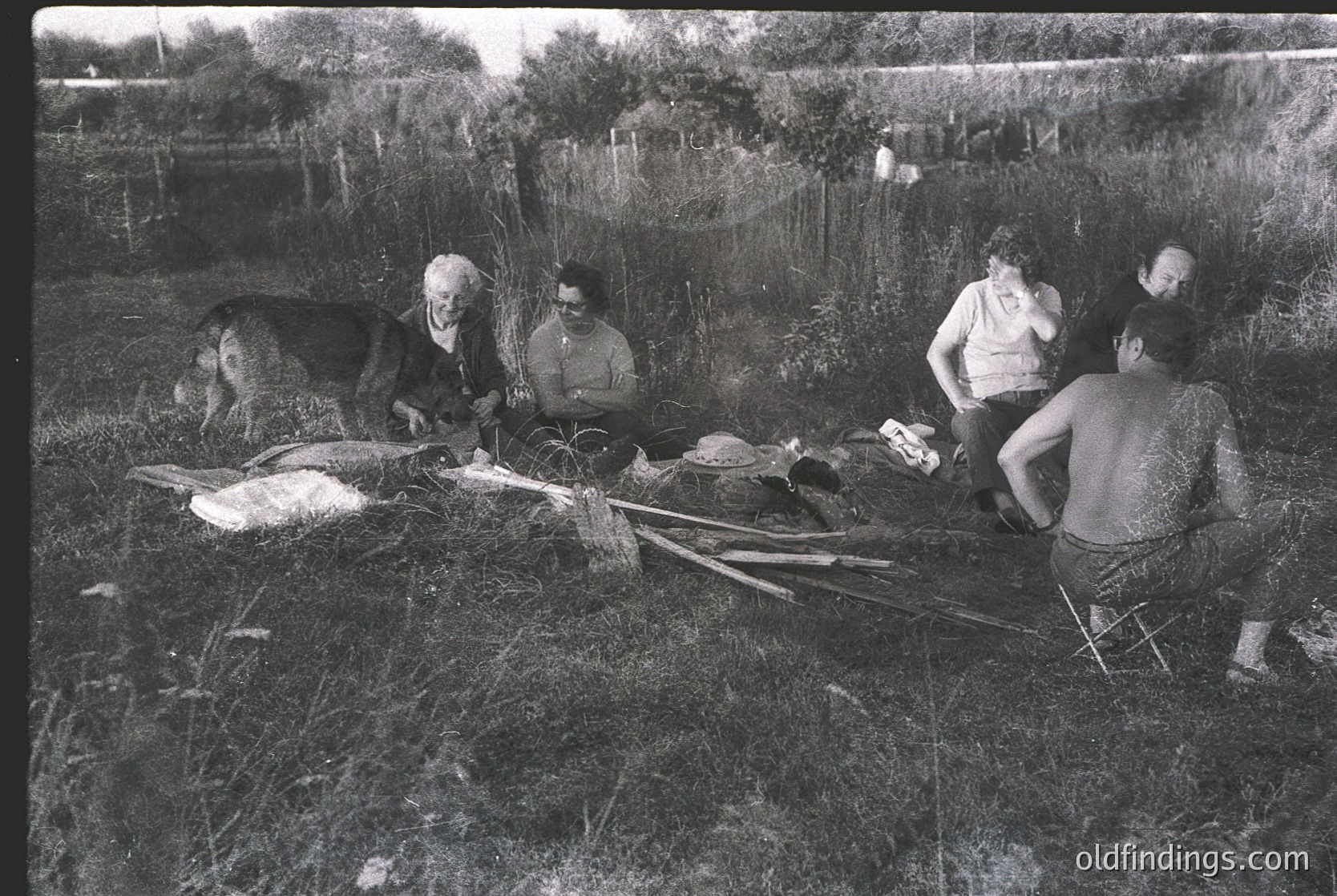 Vintage black-and-white rural gathering: group of six adults seated on grass, preparing food outdoors. Skinned animal carcass and butchering tools visible. Mid-20th century agricultural scene, likely Eastern Europe.