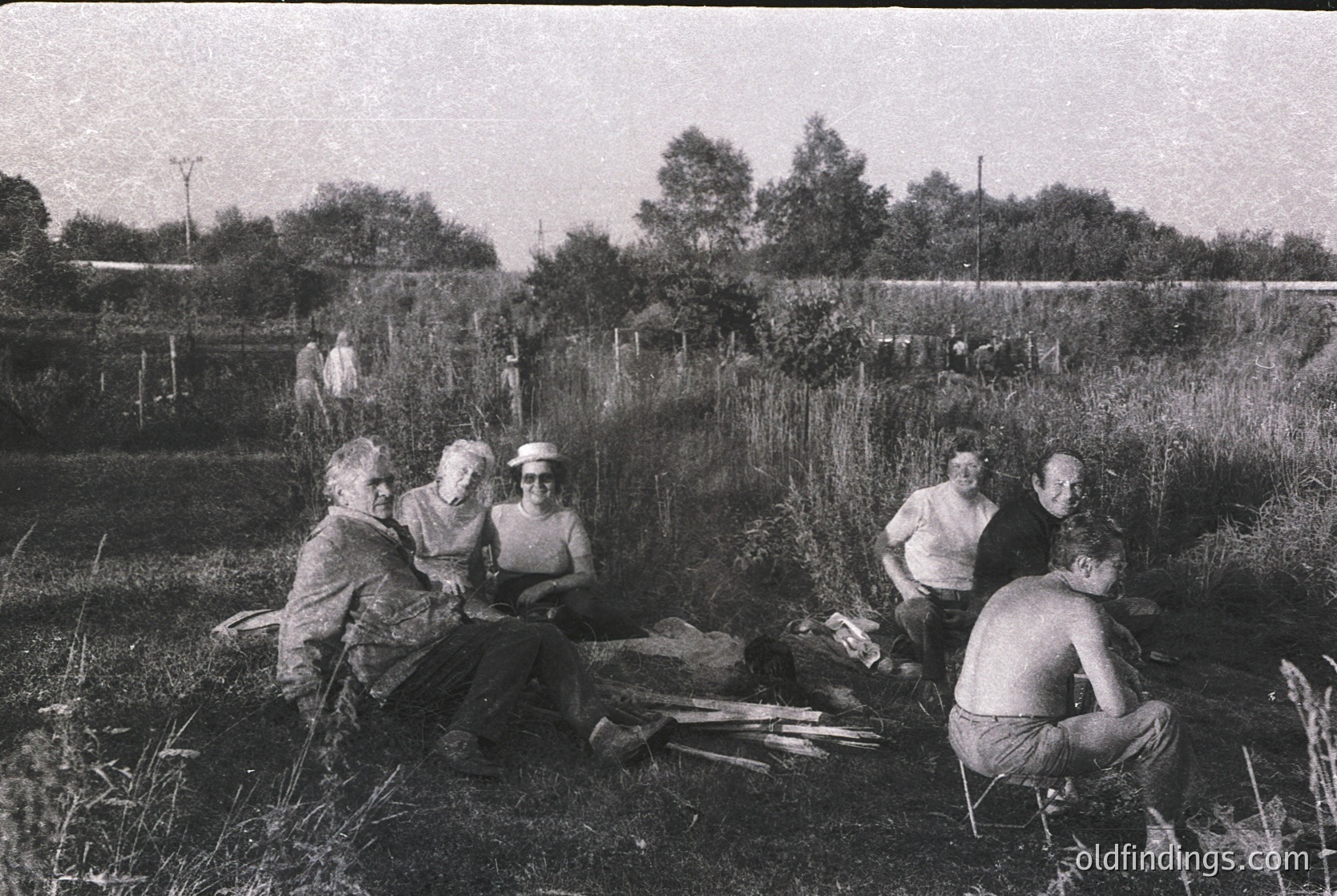Five individuals seated in rural field, mid-20th century. Group includes three women in dresses and two men in shorts, posing near wooden planks and tools. Dense vegetation and distant trees suggest agricultural or communal land use. Likely Eastern European or Soviet-era setting.