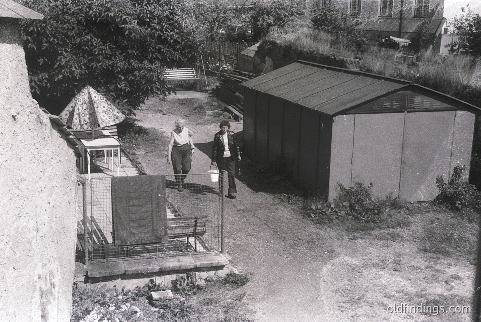 Mid-20th century backyard with two men standing near a metal-framed clothesline and a corrugated shed. Concrete steps lead to a raised platform with a wooden bench. Overgrown vegetation and a small tree frame the scene. Likely residential, Eastern European setting.