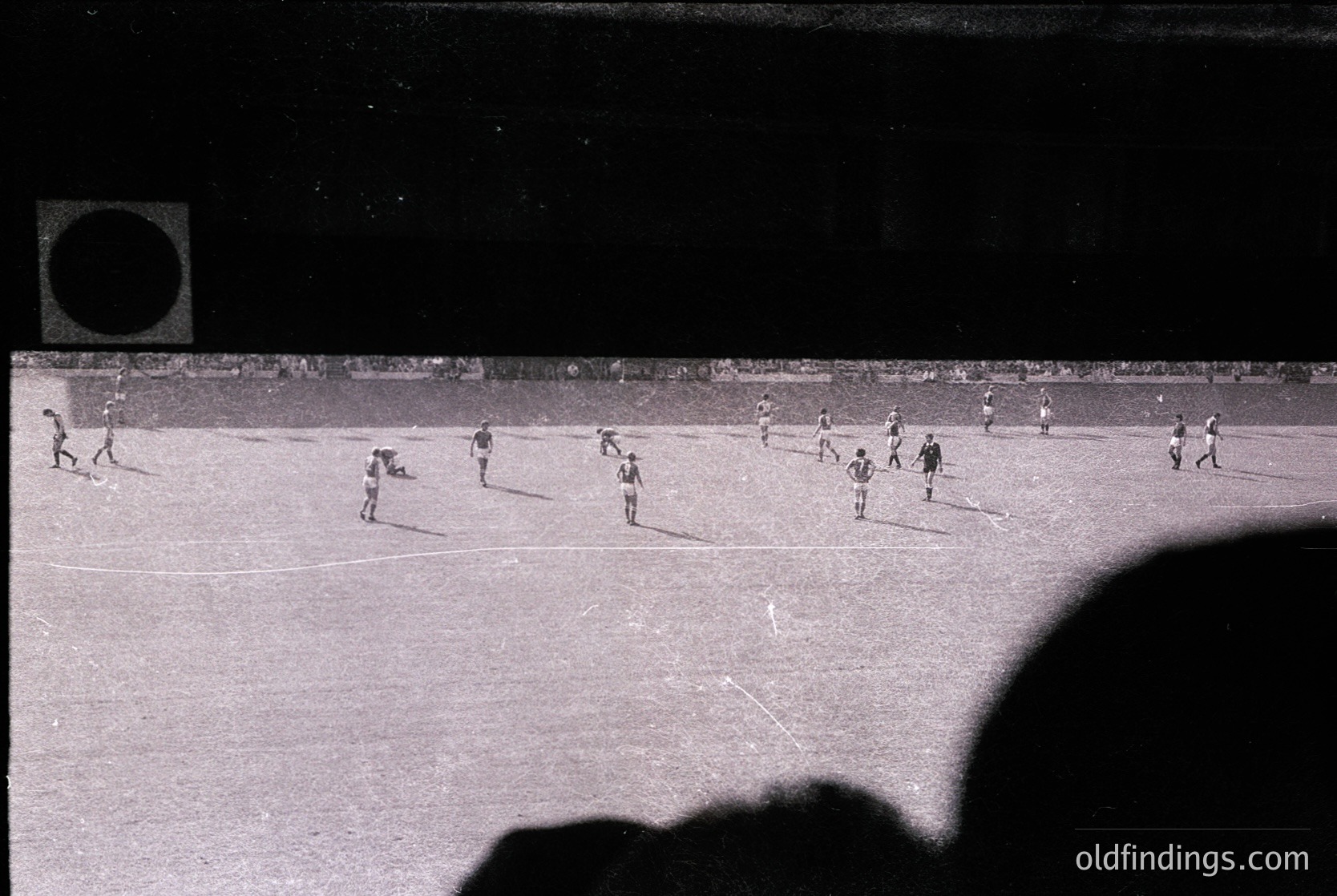 Vintage black-and-white shot of a soccer match in progress, likely mid-20th century. Players in striped jerseys and shorts engage in action on a grass field with visible goalposts. Crowd silhouettes visible in stands, suggesting a stadium setting. Dynamic composition captures intensity of the game.