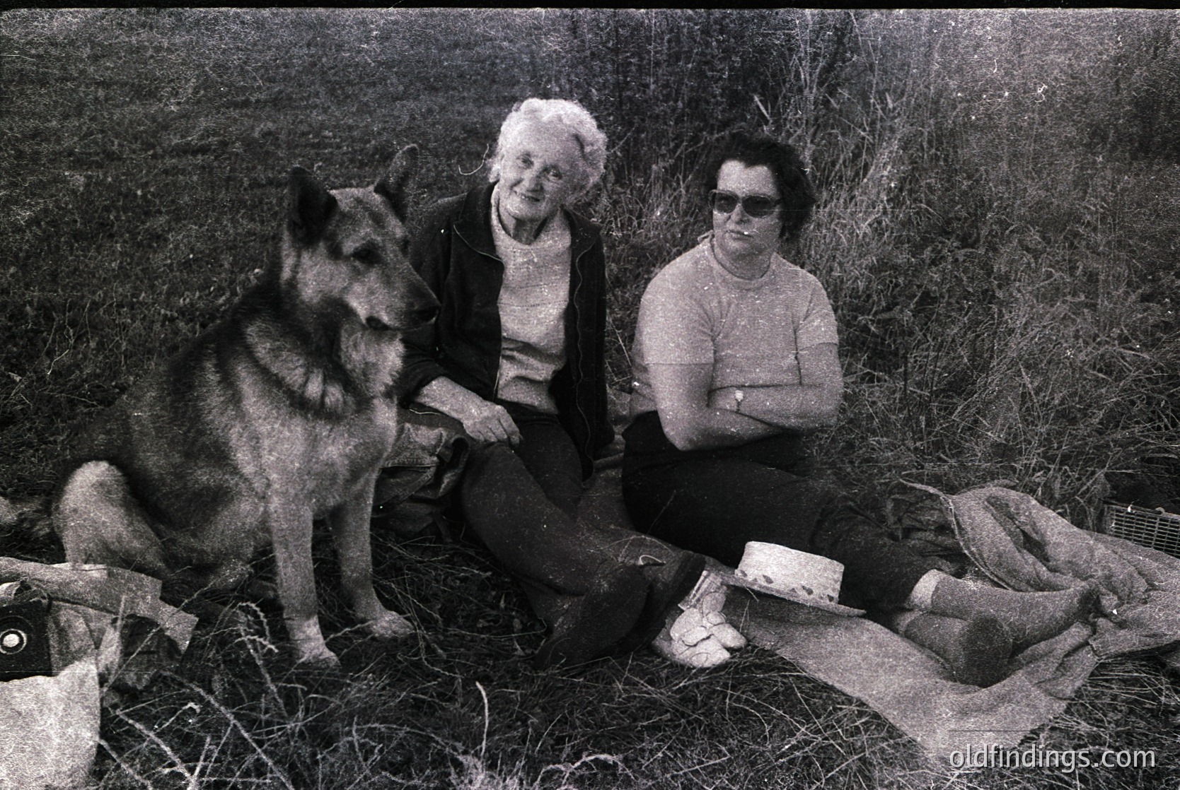 Black-and-white snapshot of two women and a German Shepherd in a rural setting, likely 1960s–1970s. Elderly woman in a cardigan and younger woman in sunglasses pose with the dog, seated on a blanket with a box and shoes nearby. Grassland background suggests countryside or farmland.