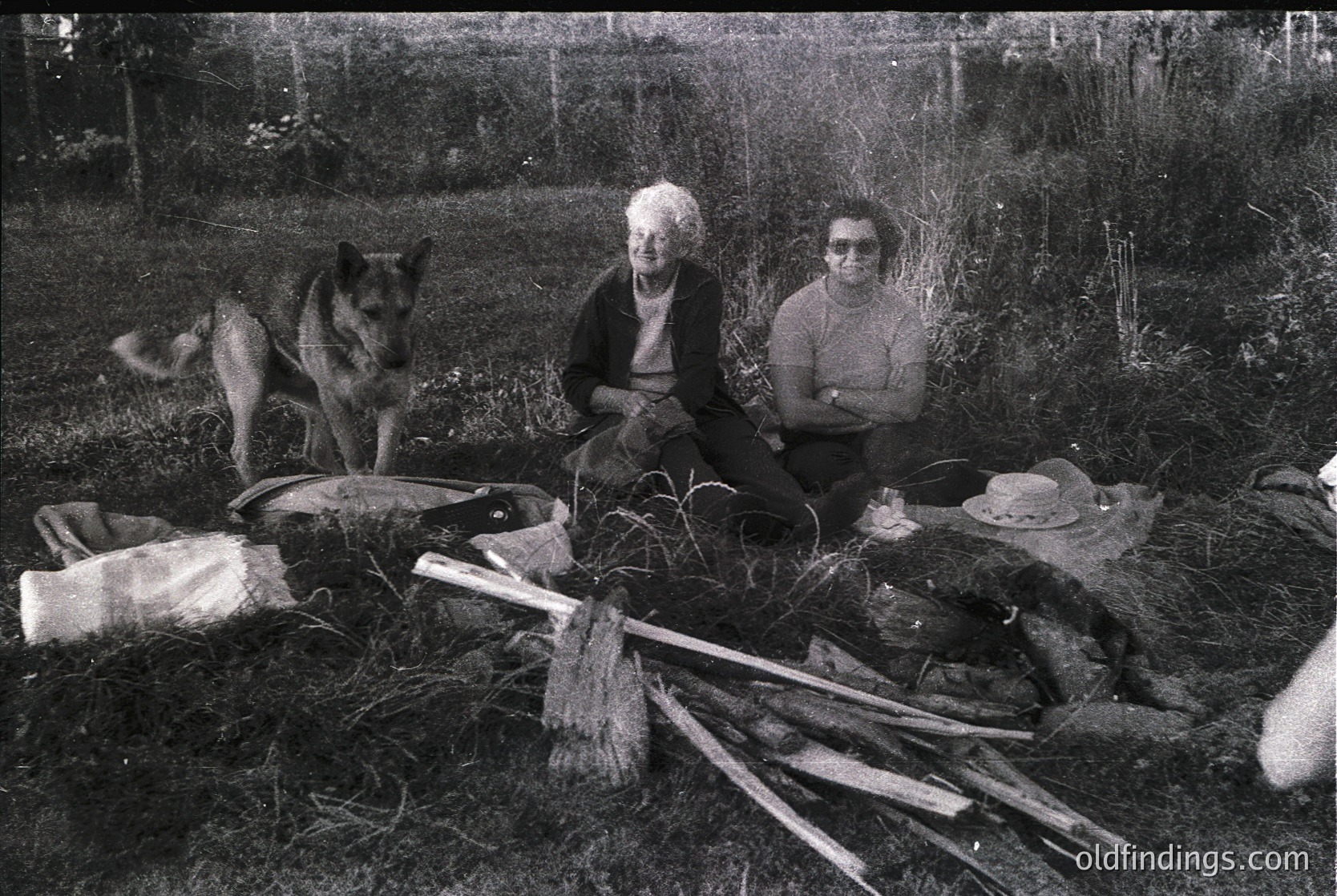 Two individuals seated outdoors on grass beside rustic tools and a dog, likely mid-20th century. The older woman wears a dark sweater, the younger a striped sweater with a hat. Surrounding items include a broom, woven basket, and wooden sticks. Rural, agricultural setting.