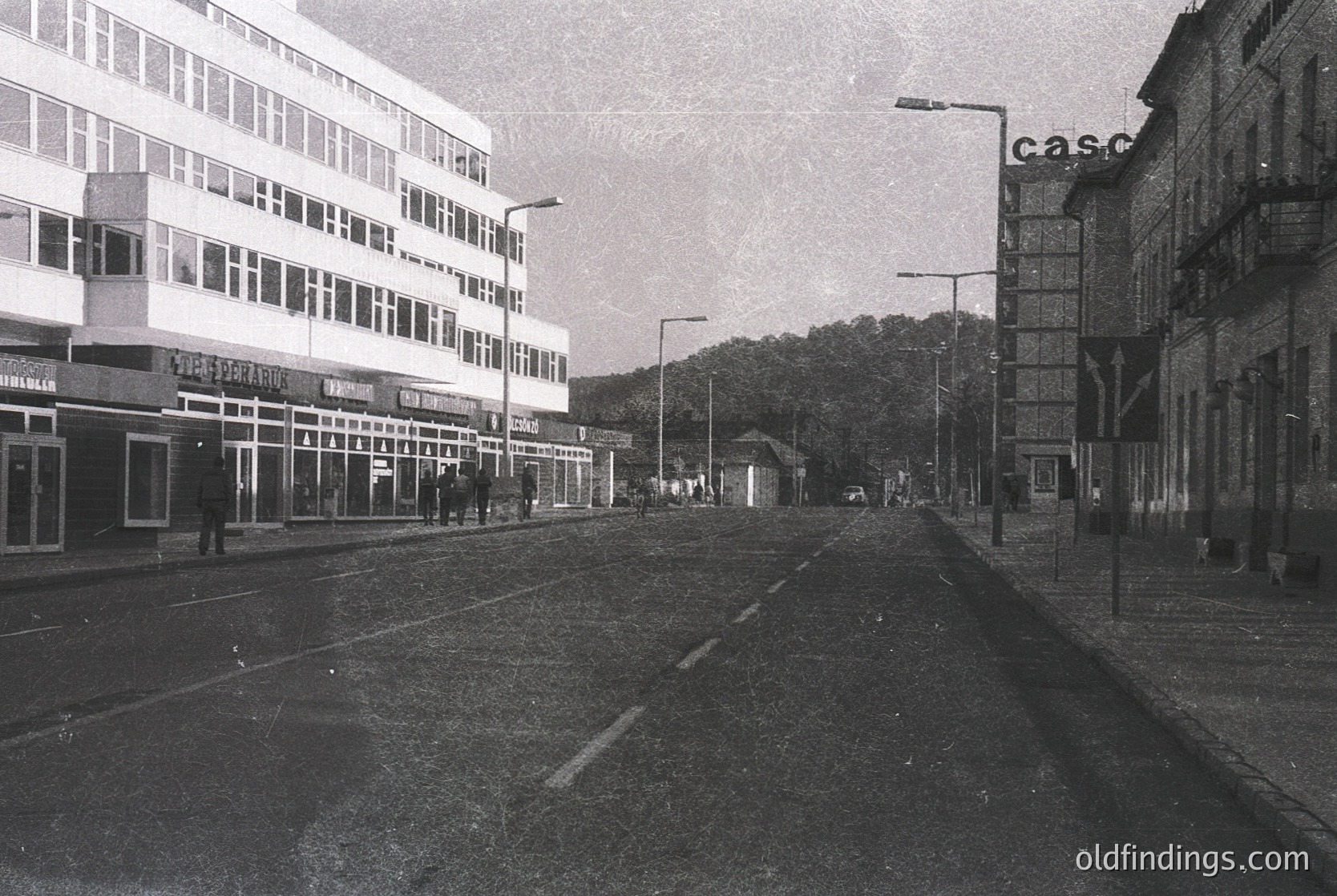 Mid-century urban street with Brutalist architecture—prominent concrete buildings featuring large windows and geometric designs. "Casco" signage suggests a European setting. Empty tram tracks and sparse pedestrians indicate a quiet, possibly early morning scene.